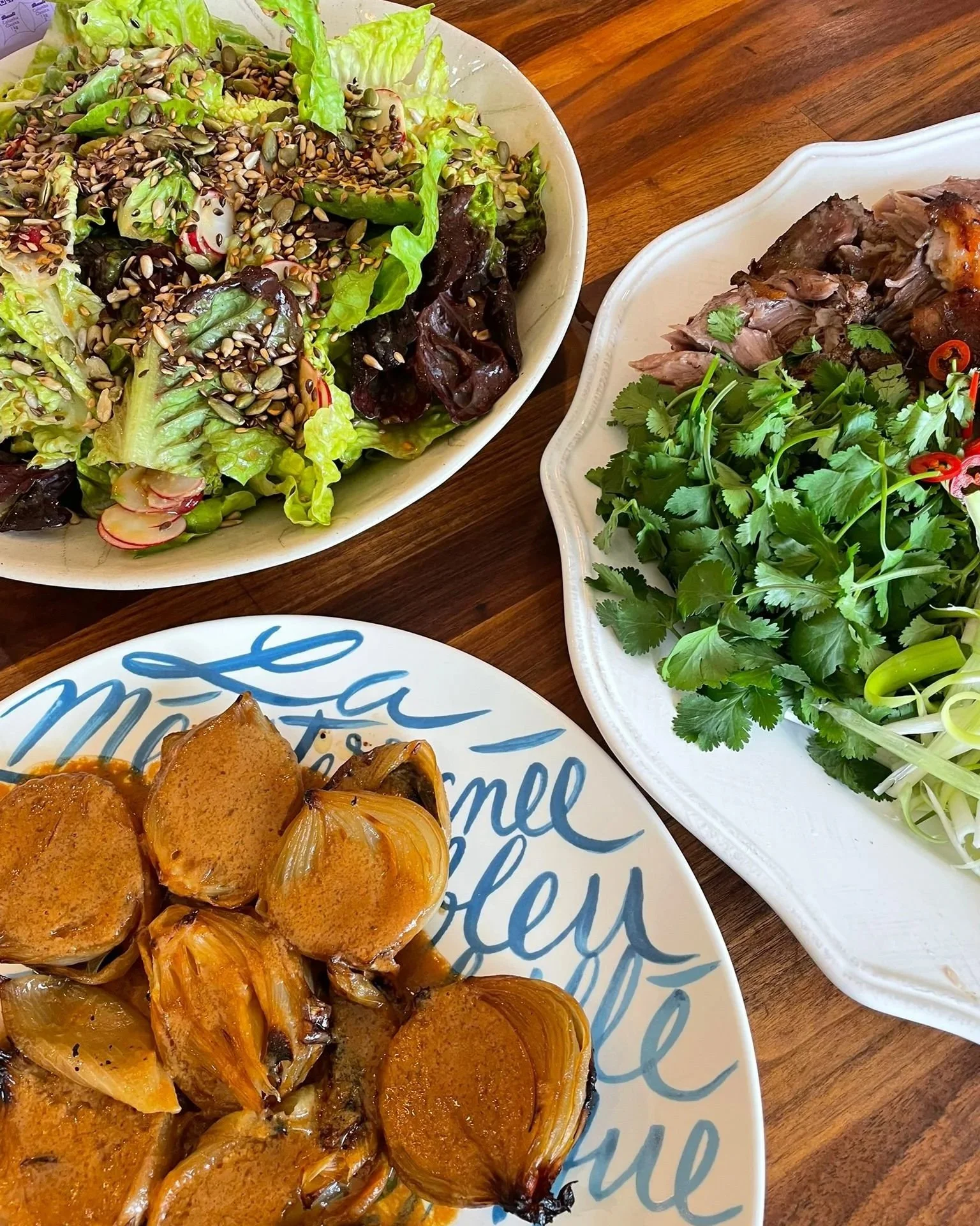 A salad with mixed greens, radishes, sunflower seeds, and a light dressing, a platter of cooked meat garnished with cilantro and red chili, and a plate of cooked garlic cloves in sauce on a wooden table.