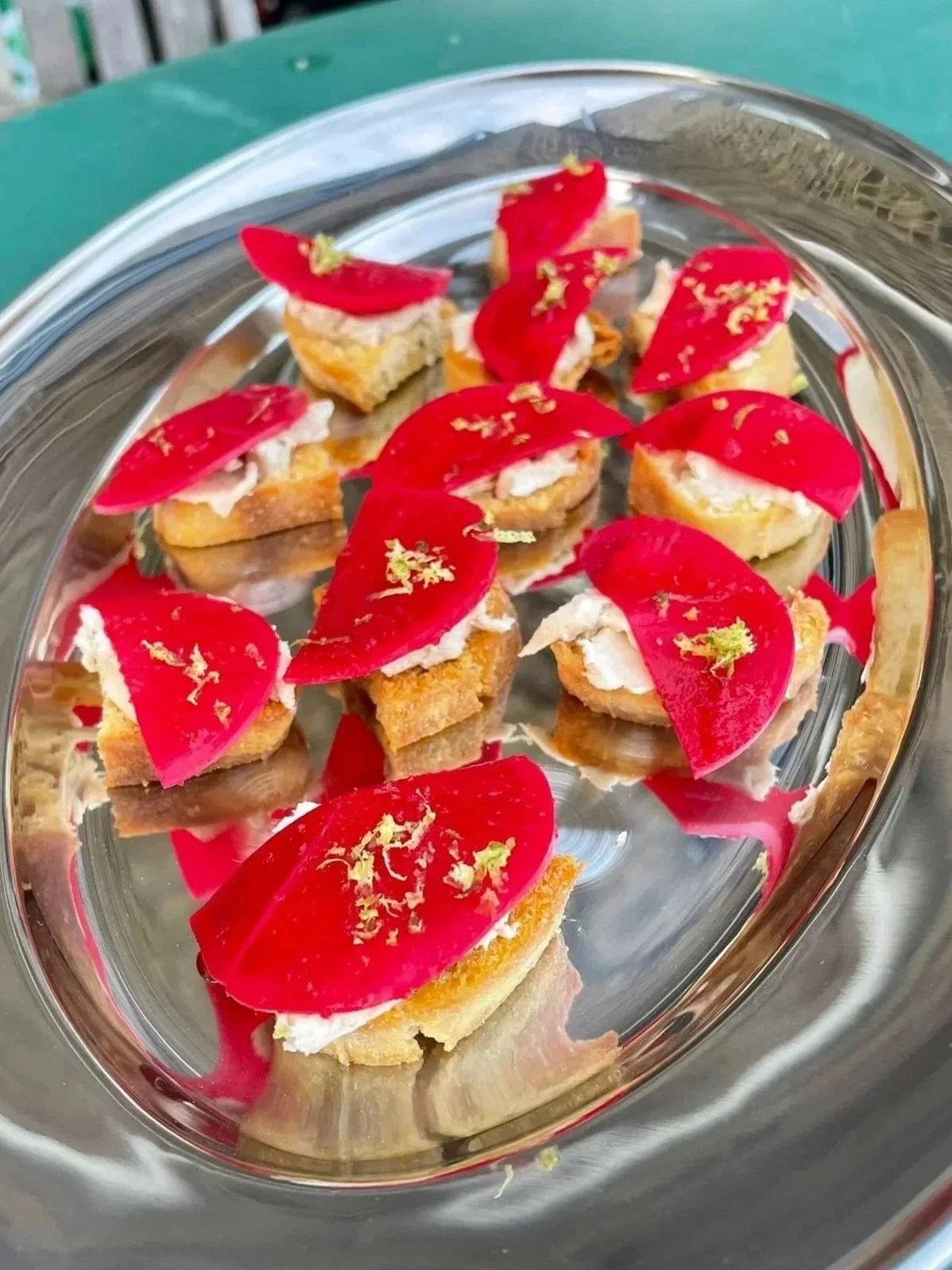 Small appetizers on a silver tray, topped with red slices of beet, cream cheese, and garnished with lemon zest.