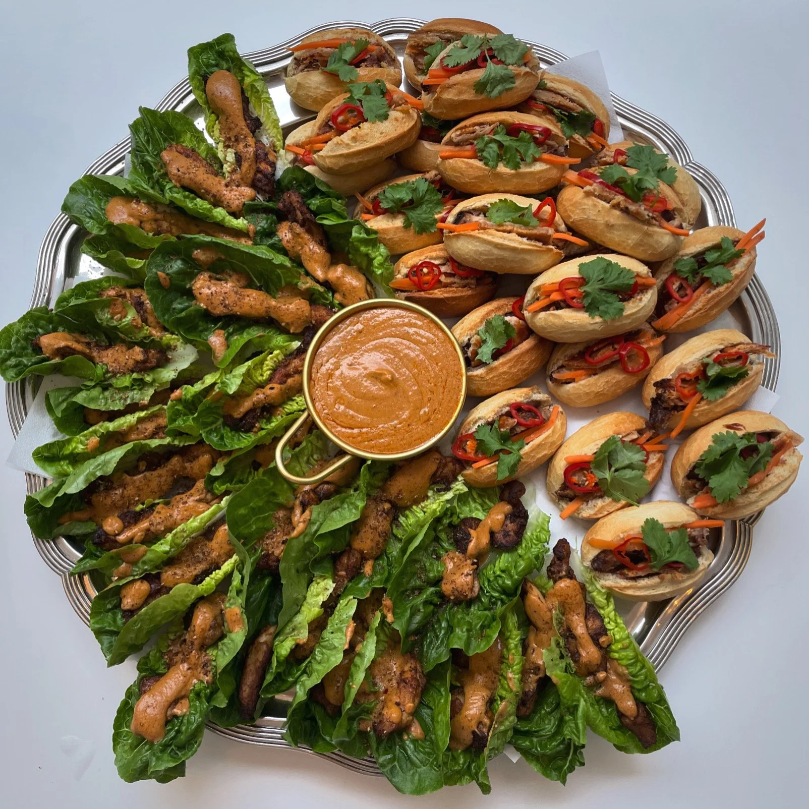 Tray of assorted Asian fusion appetizers including lettuce wraps with grilled meat, mini sandwiches with vegetables and cilantro, and a bowl of dipping sauce.