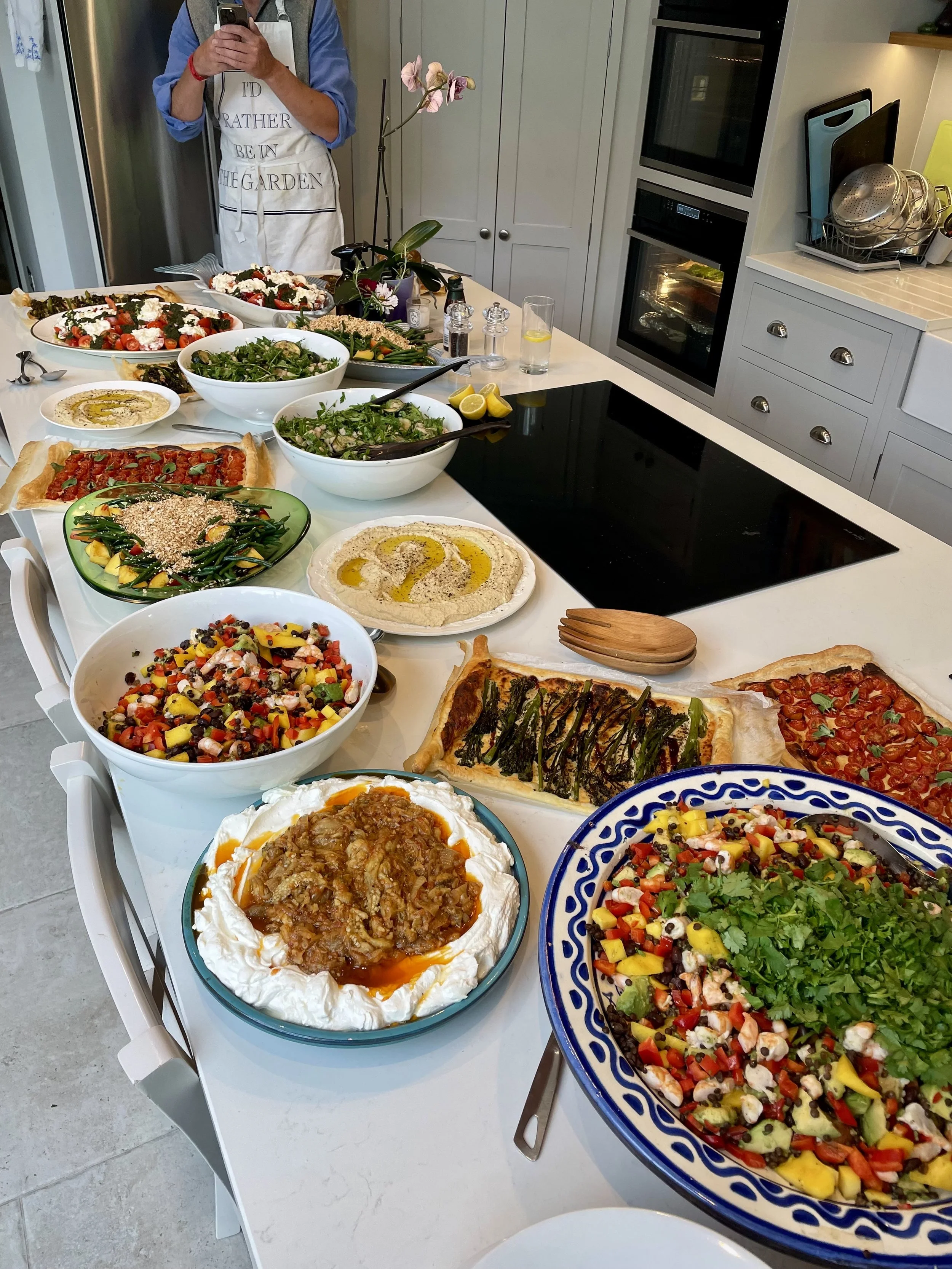 A kitchen counter filled with various dishes including salads, pizzas, hummus, and a person taking a selfie in the background.