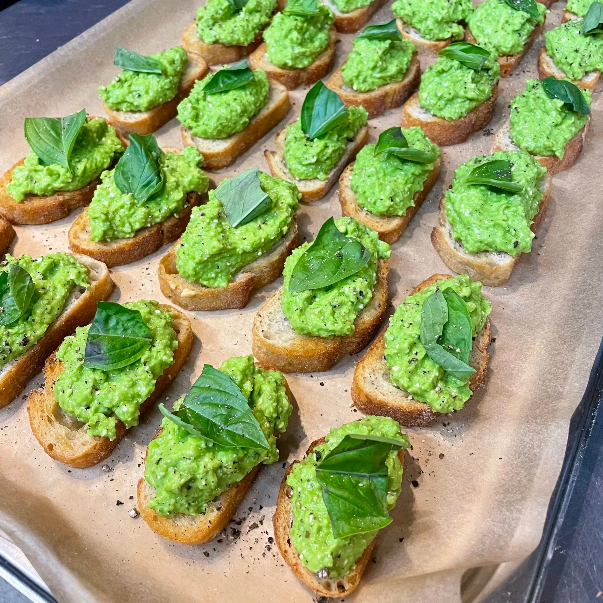 Slices of toasted baguette topped with mashed avocado, garnished with fresh basil leaves, arranged on parchment paper in a tray.