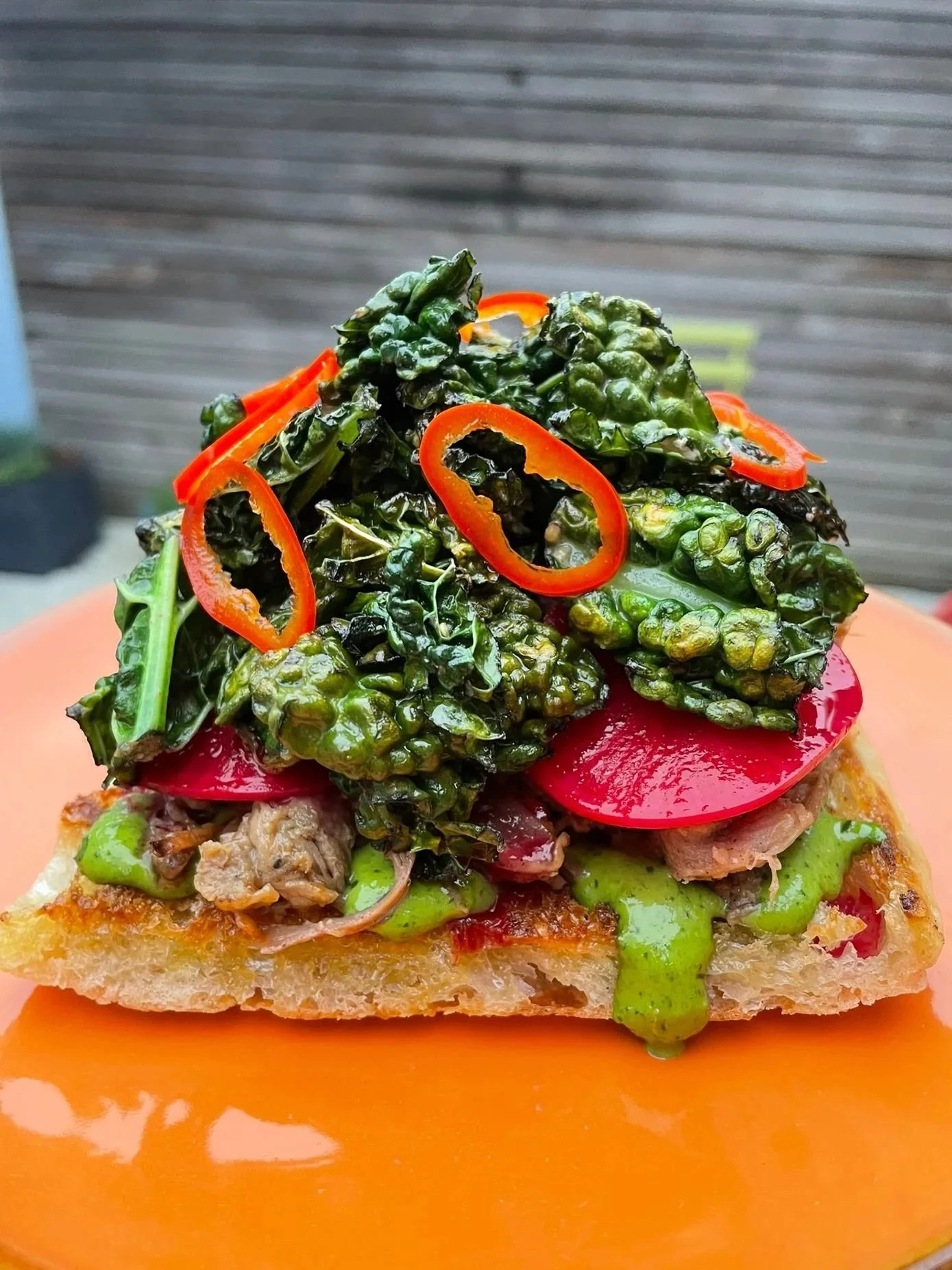 Close-up of a slice of vegetable-topped pizza with leafy greens and sliced red chili peppers, with a drizzle of green sauce and a red-orange sauce underneath.