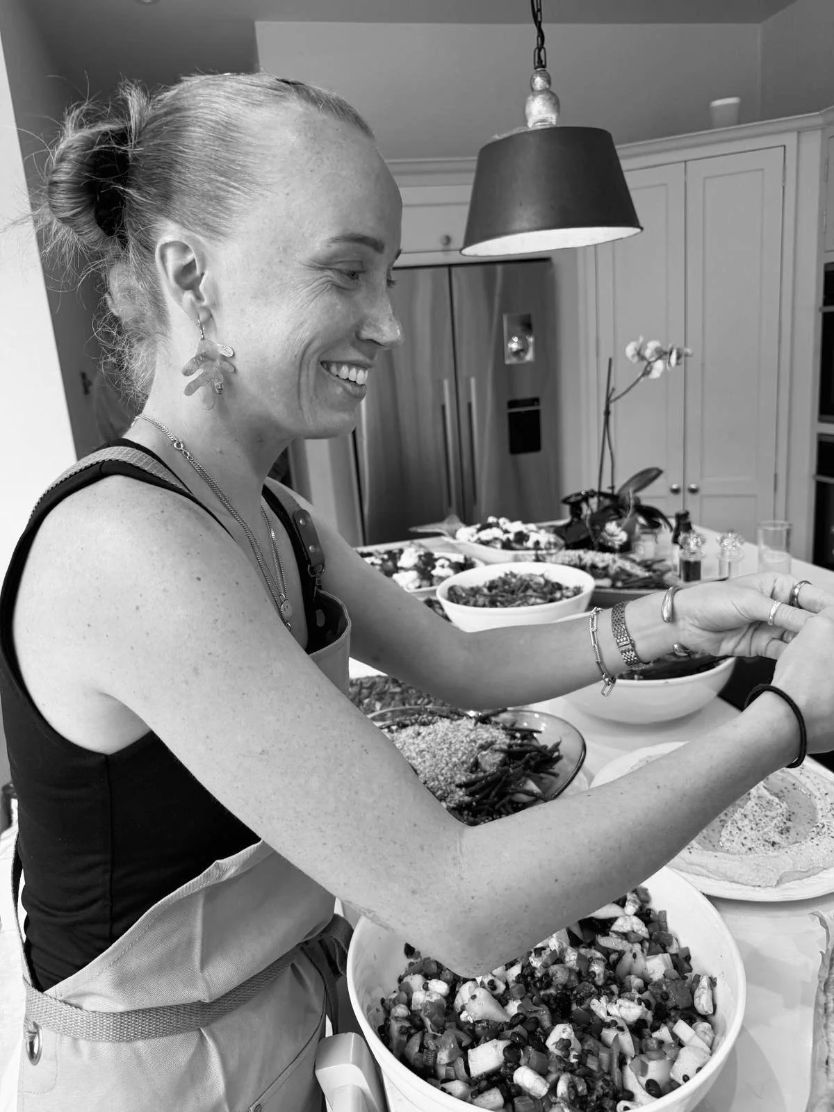 A woman with a smile, wearing a sleeveless top and jewelry, preparing food in a kitchen with bowls of salad and other dishes on the counter.