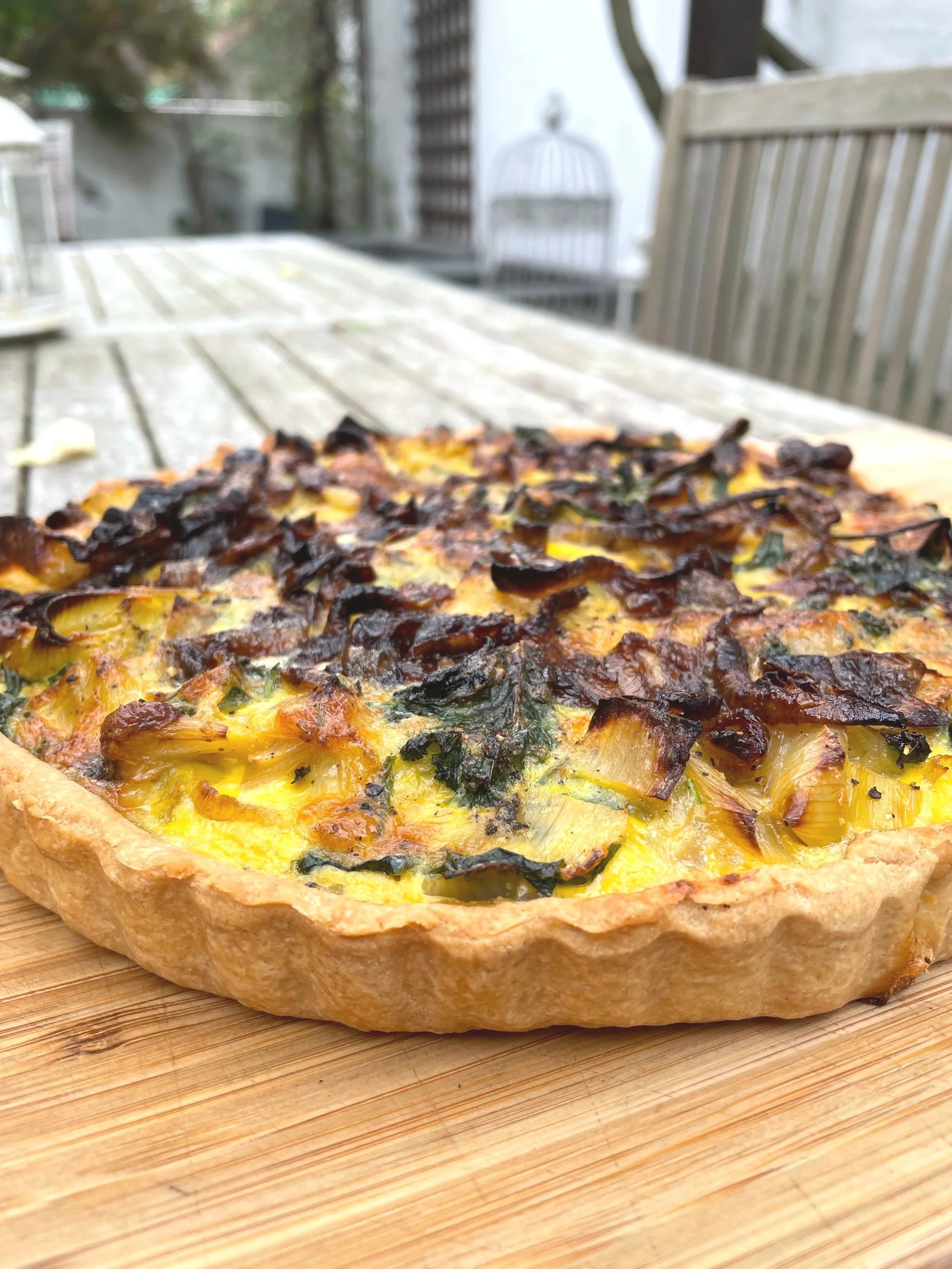 A vegetable quiche on a wooden cutting board outdoors with patio furniture in the background.