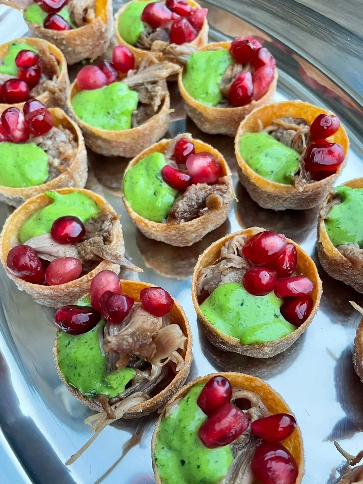 Small appetizer cups filled with shredded meat, topped with green sauce and garnished with pomegranate seeds on a silver tray.