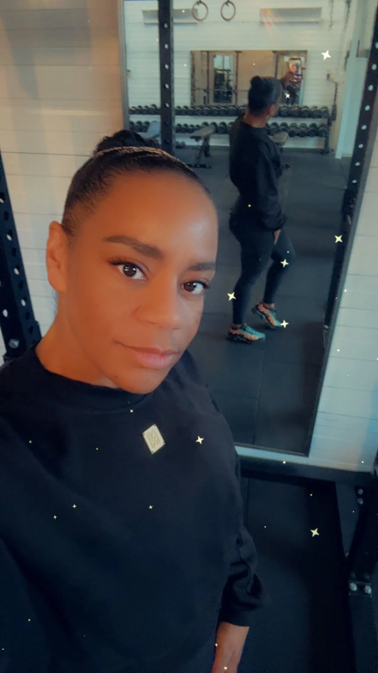 A woman taking a selfie in a gym with fitness equipment and a mirror in the background, wearing a black top, with stars and sparkles overlaying the image.