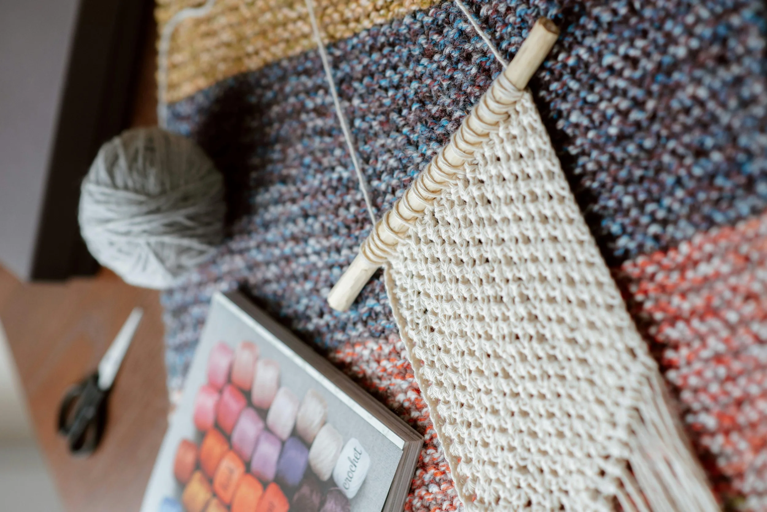 Knitting supplies including a ball of white yarn, a knitting needle, a knitted fabric, a pair of scissors, and a color chart, all arranged on a multicolored woven surface.