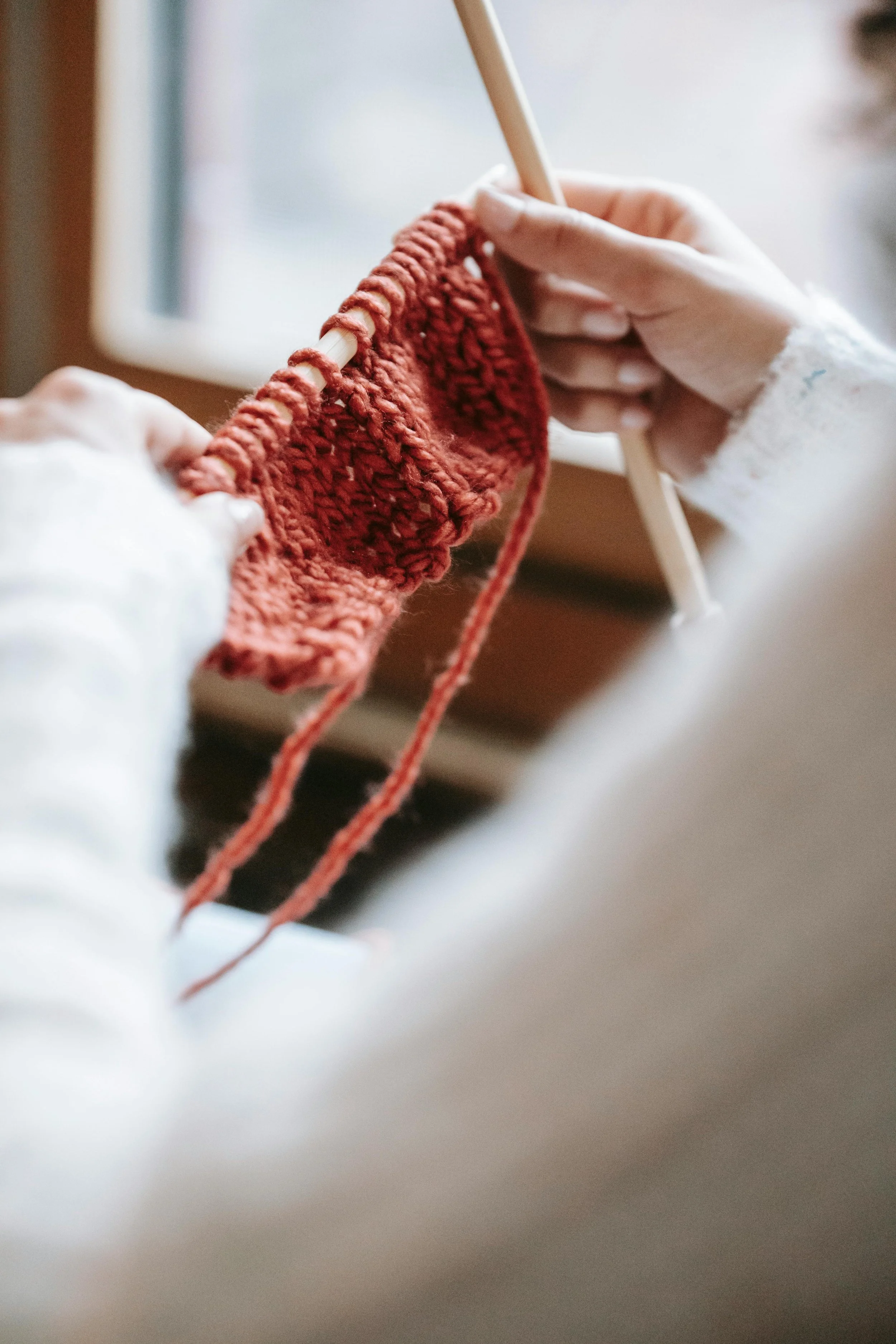 Close-up of hands knitting a piece of fabric with reddish yarn on knitting needles near a window.