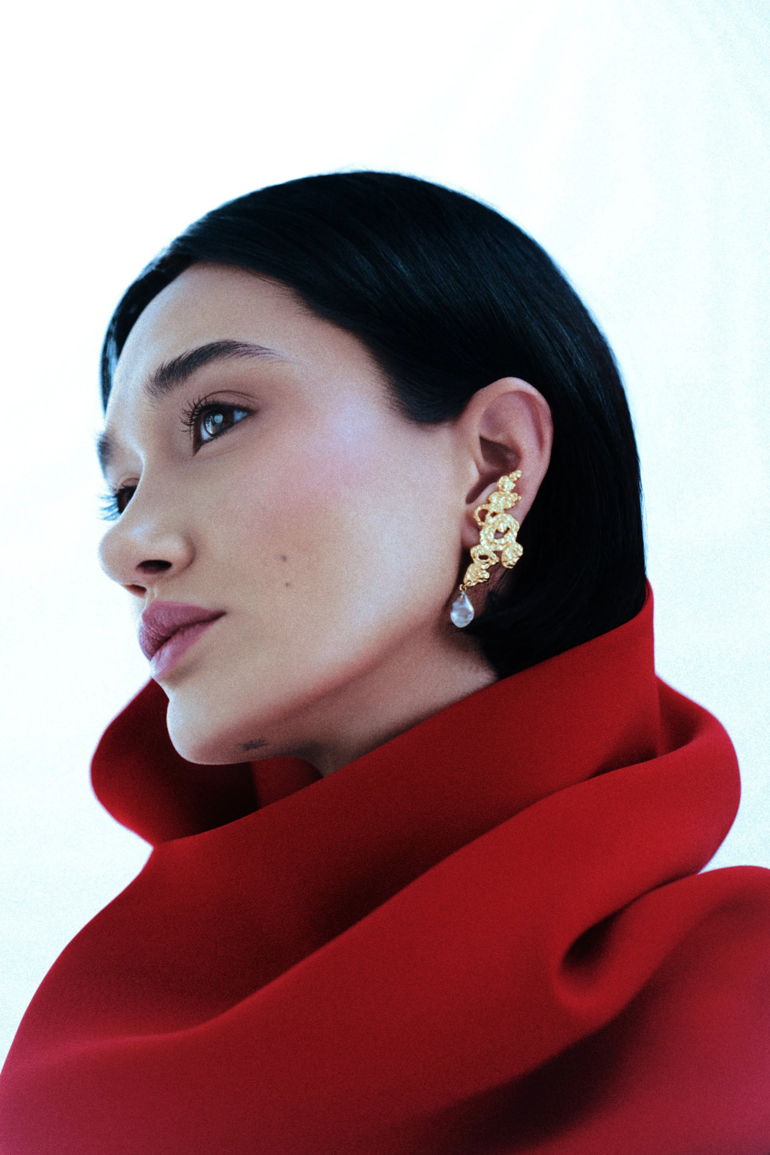 Close-up of a woman with short black hair, wearing gold earrings with dangling pearls, and a red high-collared garment against a plain white background.