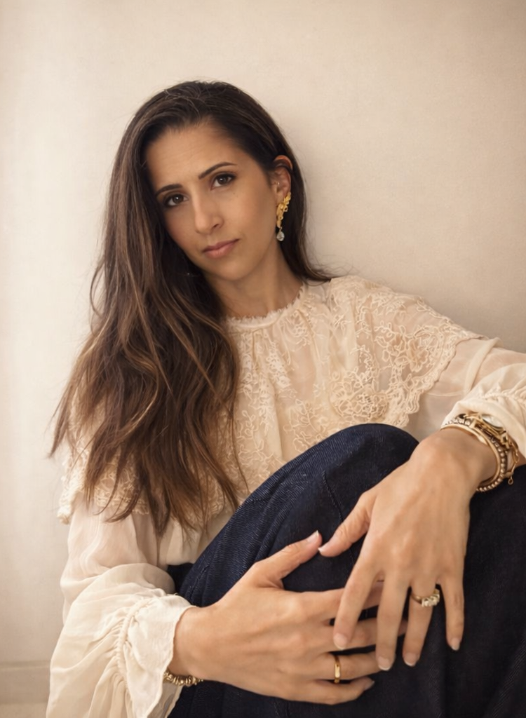 Personal Fashion Stylist .A woman with long brown hair wearing a cream lace blouse and gold jewelry, sitting with her arm resting on her knee.