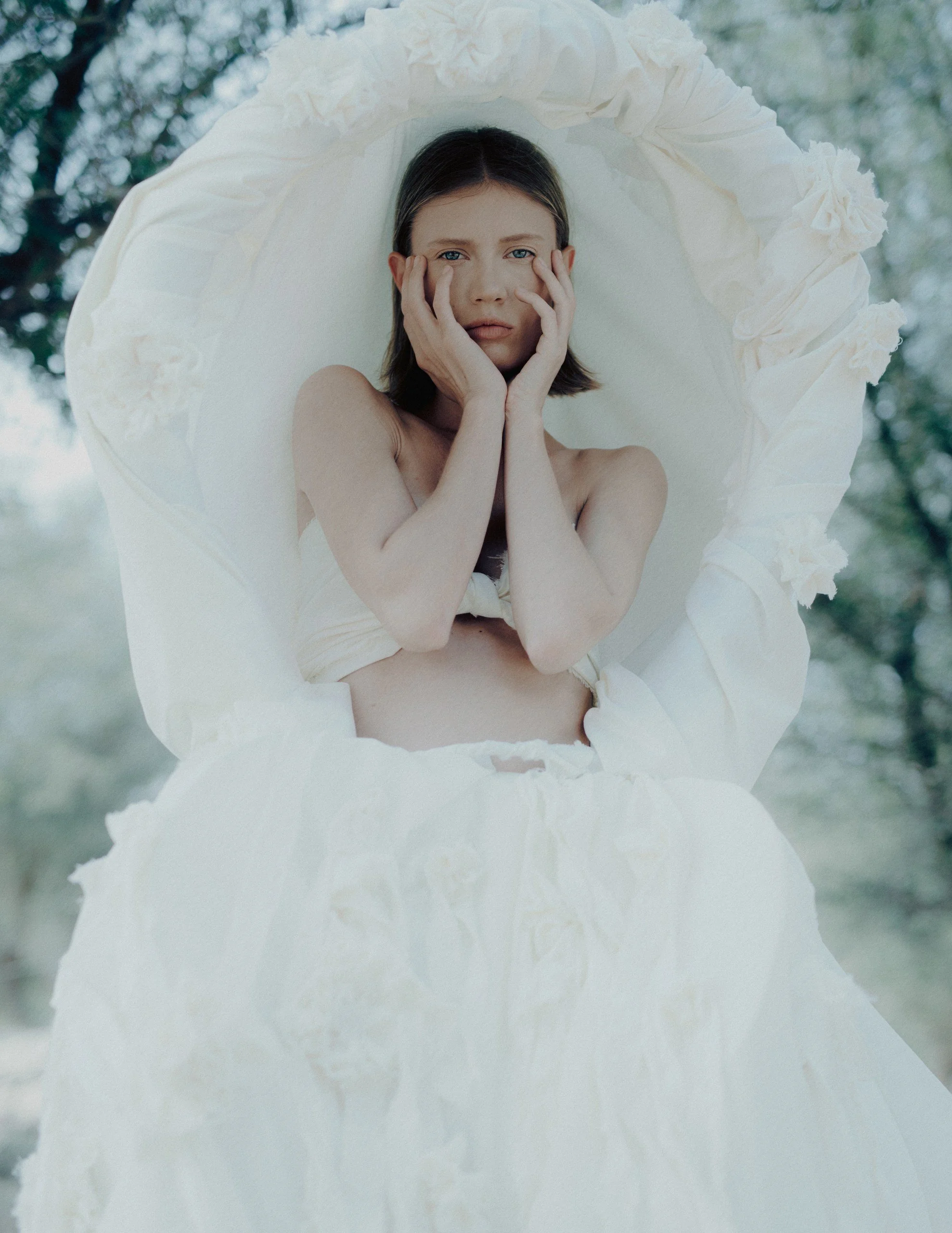 A woman with dark hair and blue eyes holding her face with both hands, standing inside a large white flower or fabric structure outdoors.