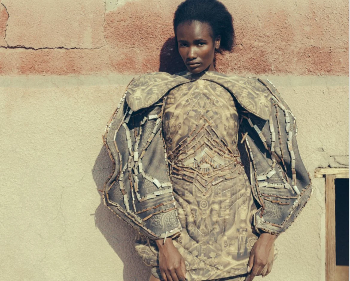 A woman standing against a wall wearing a garment made of metallic and patterned objects resembling armor.