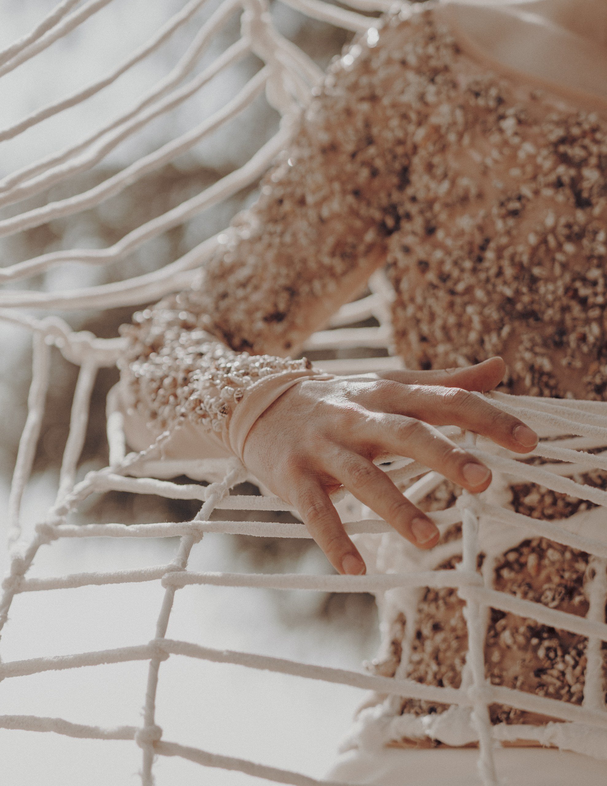 A person's hand with long fingernails resting on a white wireframe structure with textured clothing or fabric partially visible.
