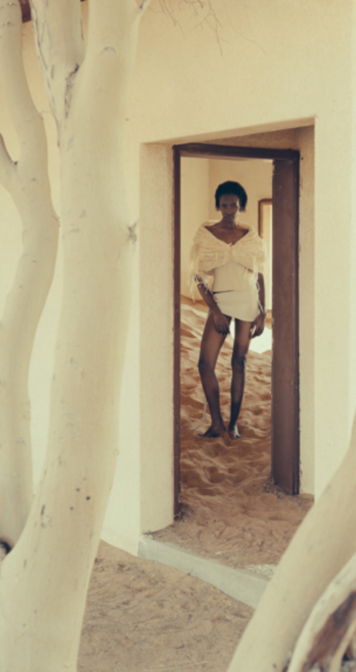Woman standing in a doorway, wearing a white top, black stockings, and shorts, with a desert or sandy floor and a tree trunk visible in the foreground.
