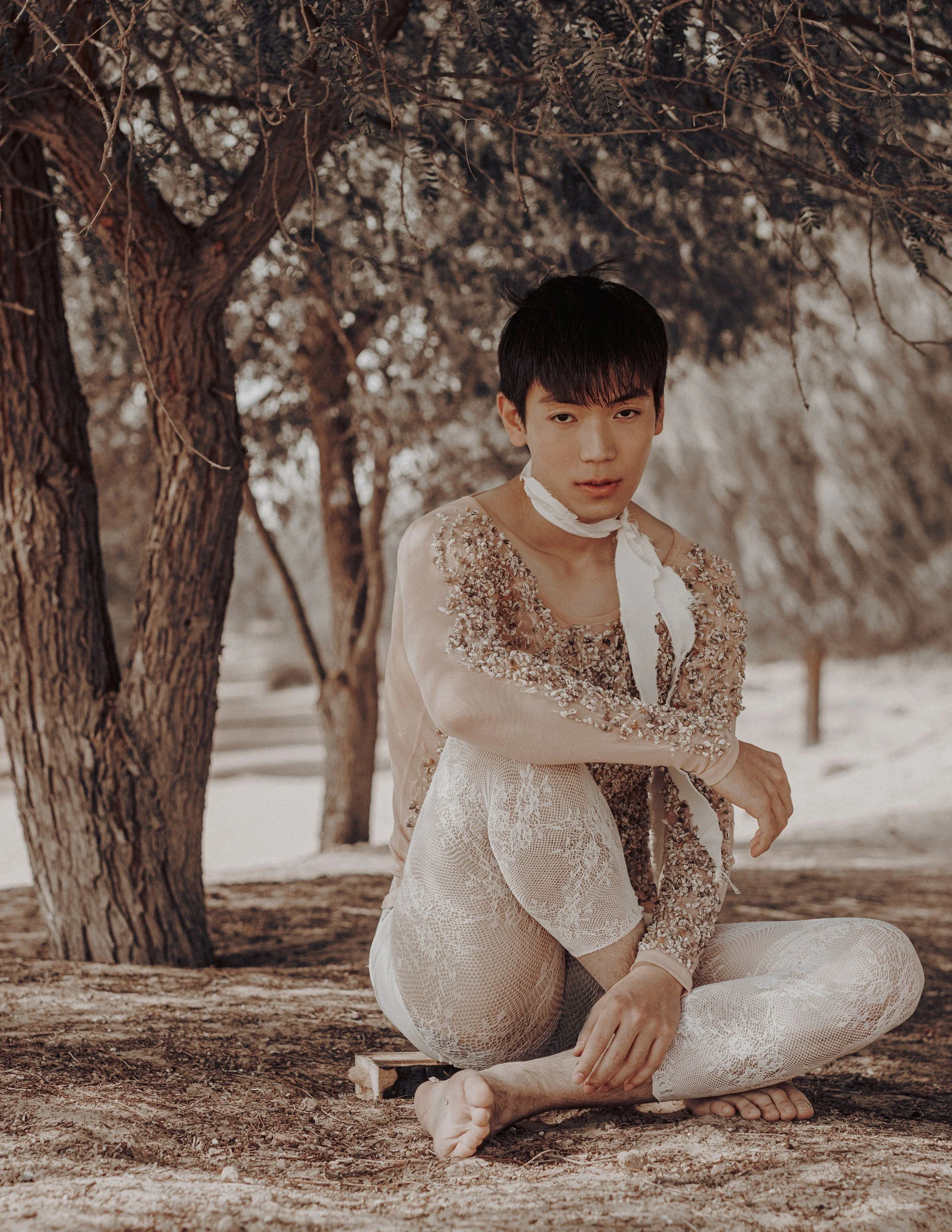 A young man with black hair sitting cross-legged on the ground outdoors. He is wearing cream-colored lace pants and a beige, embroidered jacket with floral details. A white cloth is tied around his neck. The background shows trees with brown leaves a