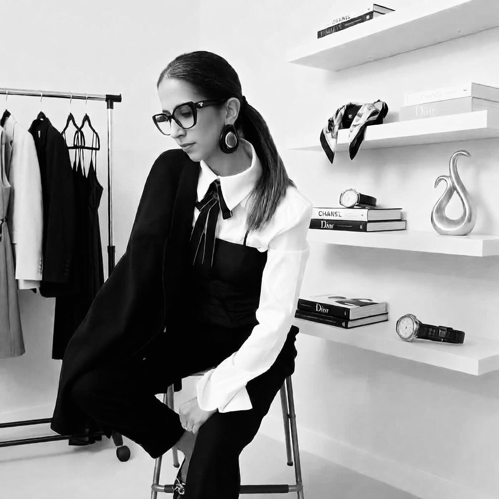 Personal fashion stylist is sitting on a stool in a modern room. She is wearing a black and white outfit with a bow tie. Behind her are shelves with books, a sculpture, and a scarf. To her left, there is a clothing rack with various dresses and tops.