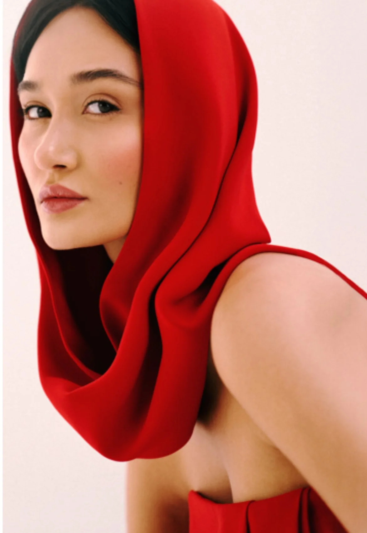 A woman with fair skin and dark hair wearing a red hooded garment that drapes over her shoulder, against a plain background.