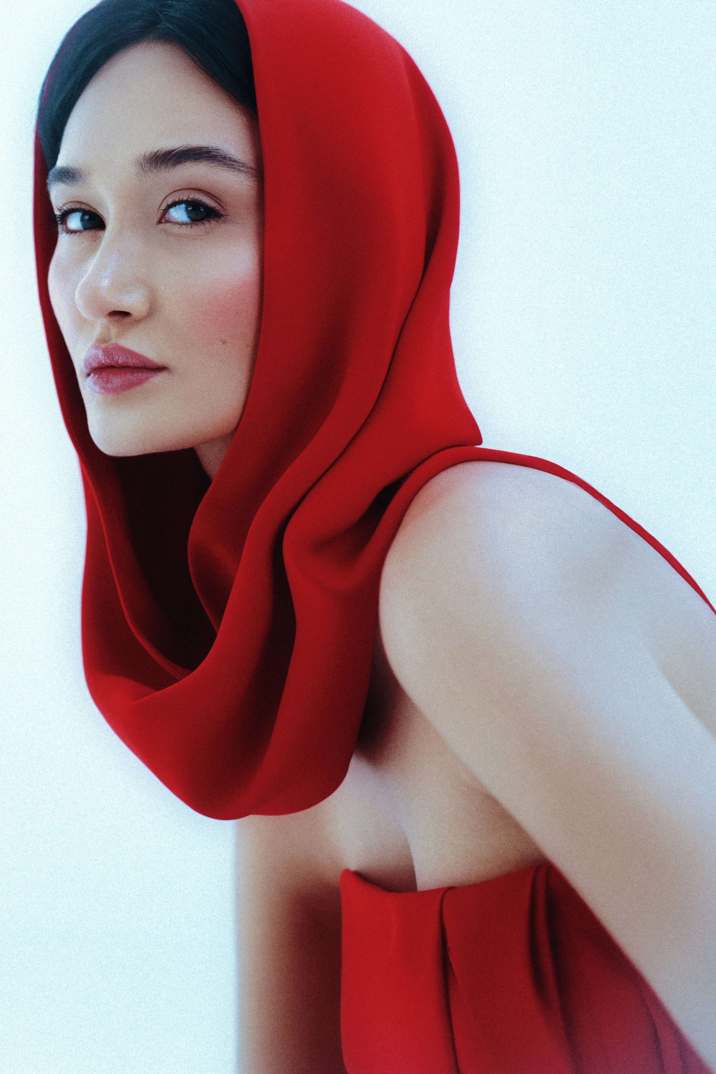 Commercial styling - A woman with fair skin and dark hair wears a red hooded garment, looking over her shoulder with a neutral expression, against a plain light background.