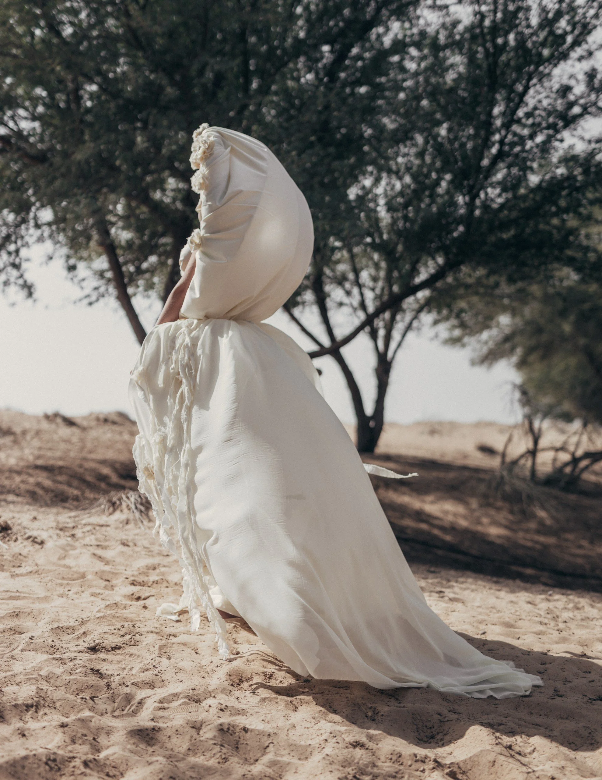A person dressed in a long, flowing white dress with a hood, standing on sandy terrain with trees in the background.