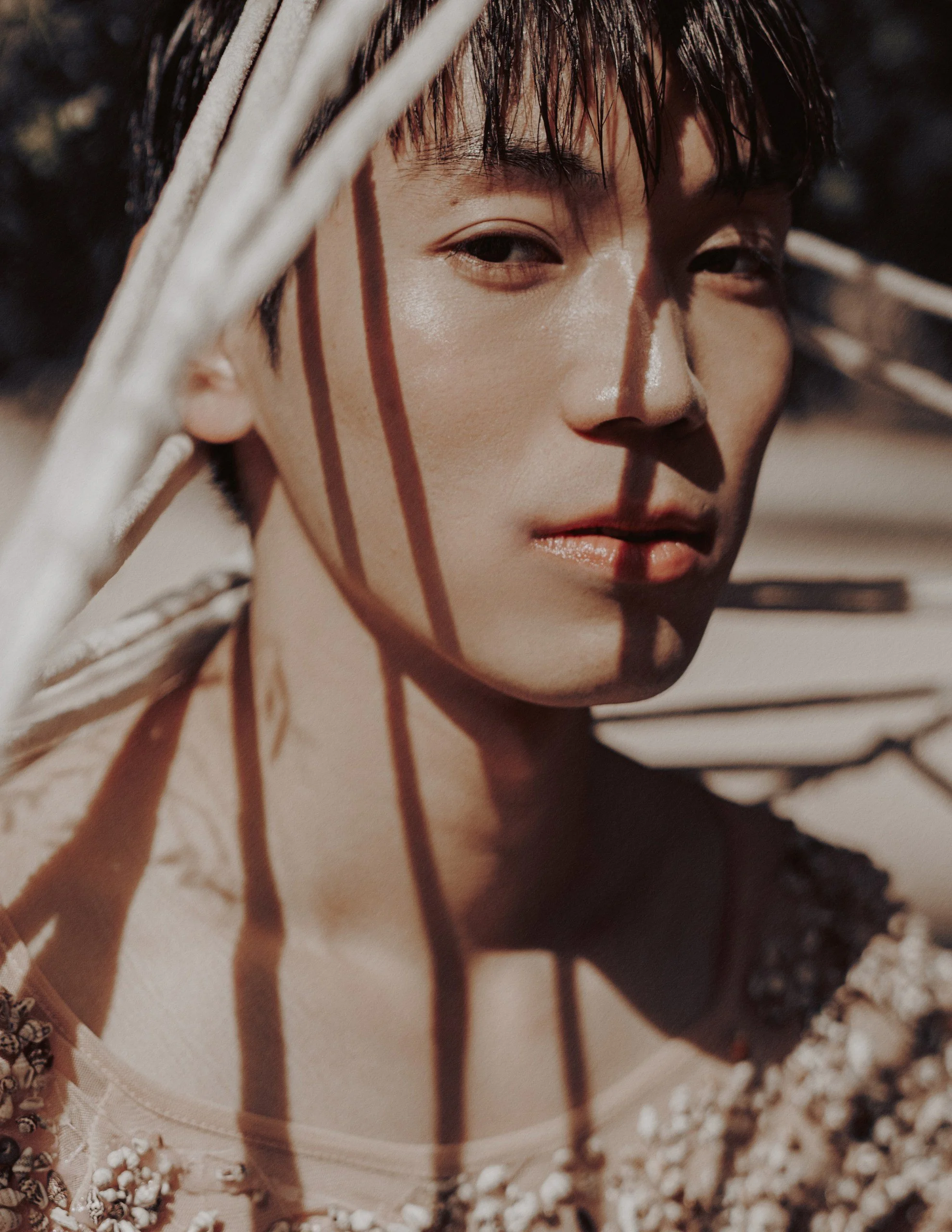 A close-up of a young person with short dark hair, looking at the camera, with shadows cast across their face from branches or leaves, in natural outdoor lighting.