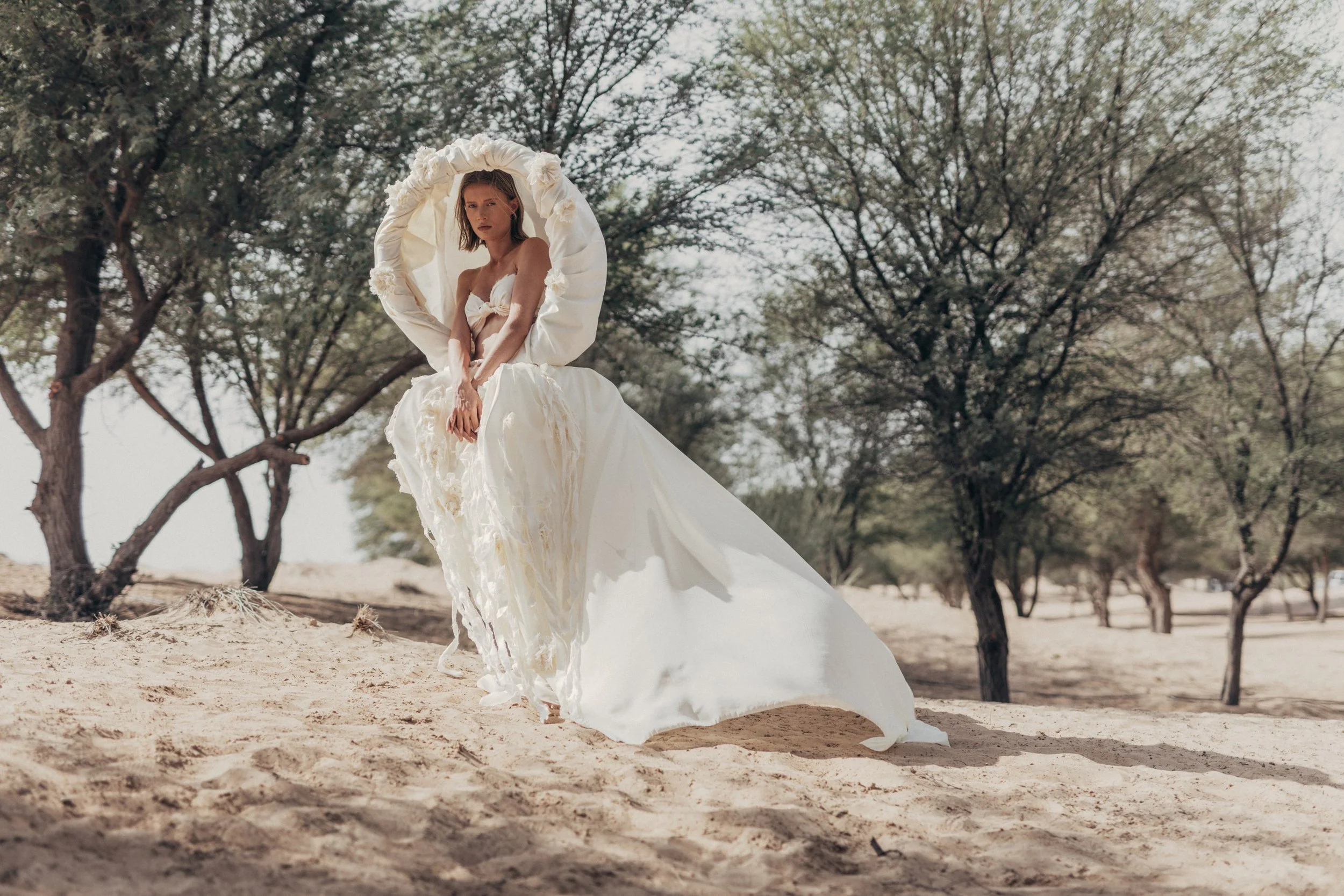 A woman wearing a white, flowing dress with a large ruffled collar stands on sandy ground among trees in a natural outdoor setting.