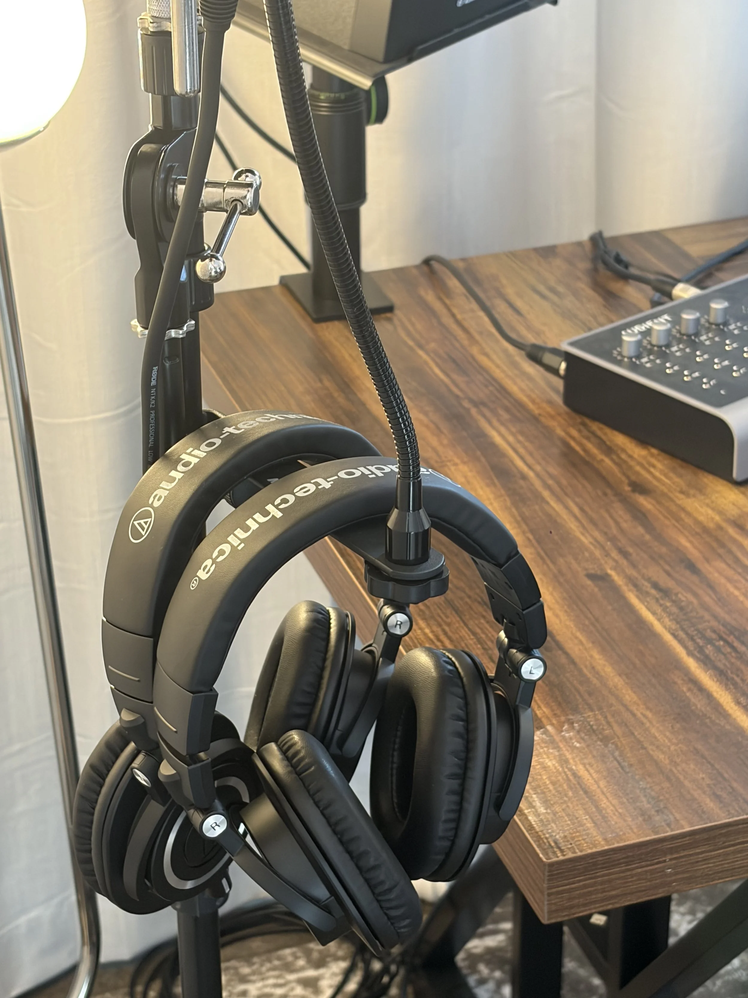 A pair of black over-ear headphones hanging on a stand with adjustable arms, positioned next to a wooden table with electronic audio equipment on it.