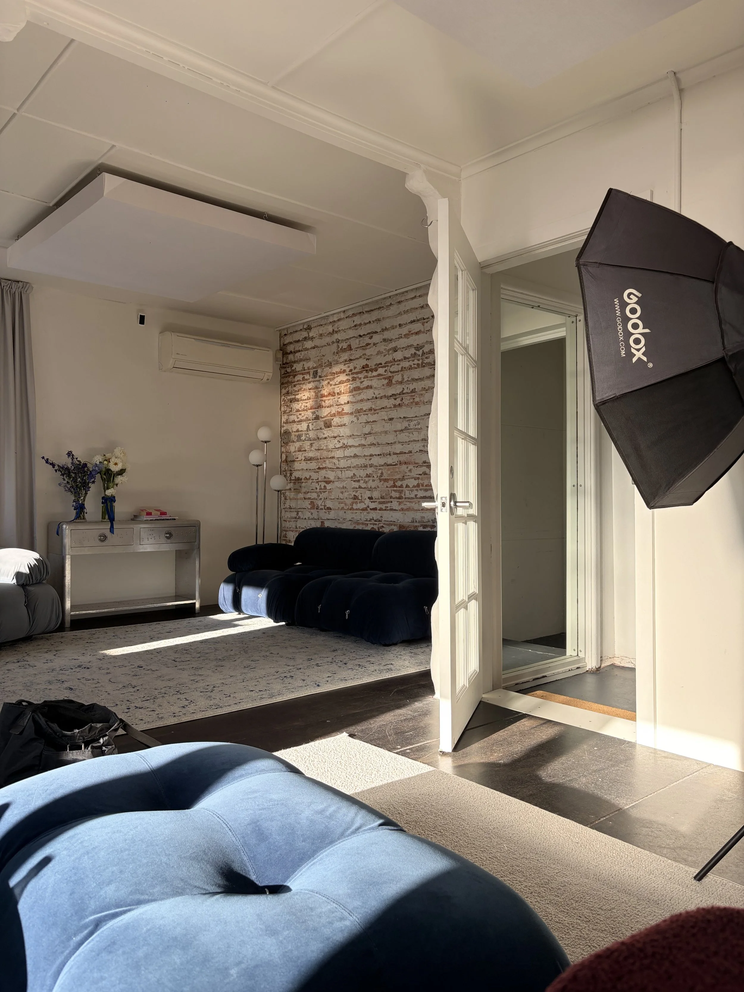 Interior of a living room with a couch, a coffee table, a vintage-style white console, and a brick wall in the background, with a photography lighting setup.