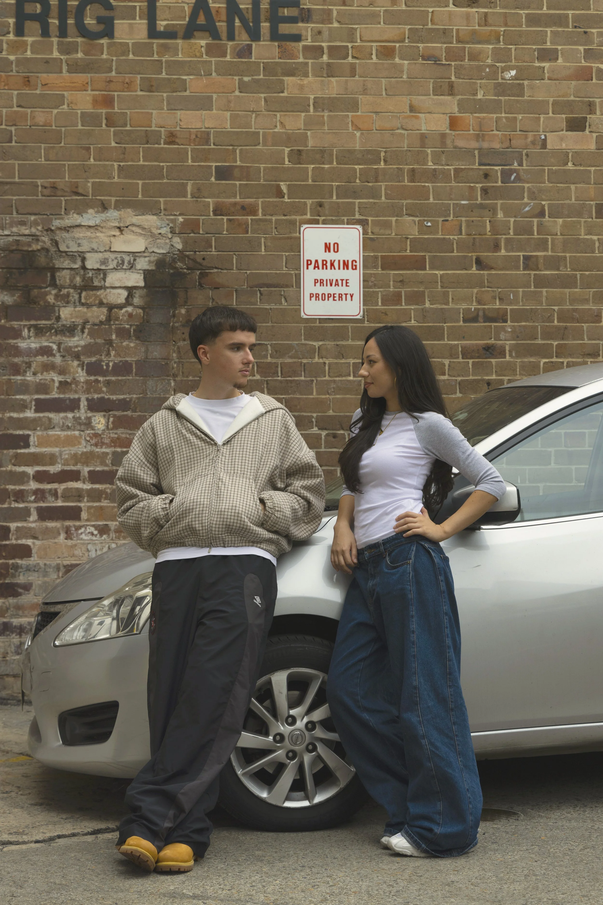 A young man and woman stand in front of a silver car, leaning against it in an alleyway with a brick wall behind them. The brick wall has a sign that reads 'No Parking Private Property.' The young man is wearing a light hoodie, black pants, and yellow boots, while the young woman is dressed in a white and gray long-sleeve shirt and wide-leg blue jeans. They are looking at each other.