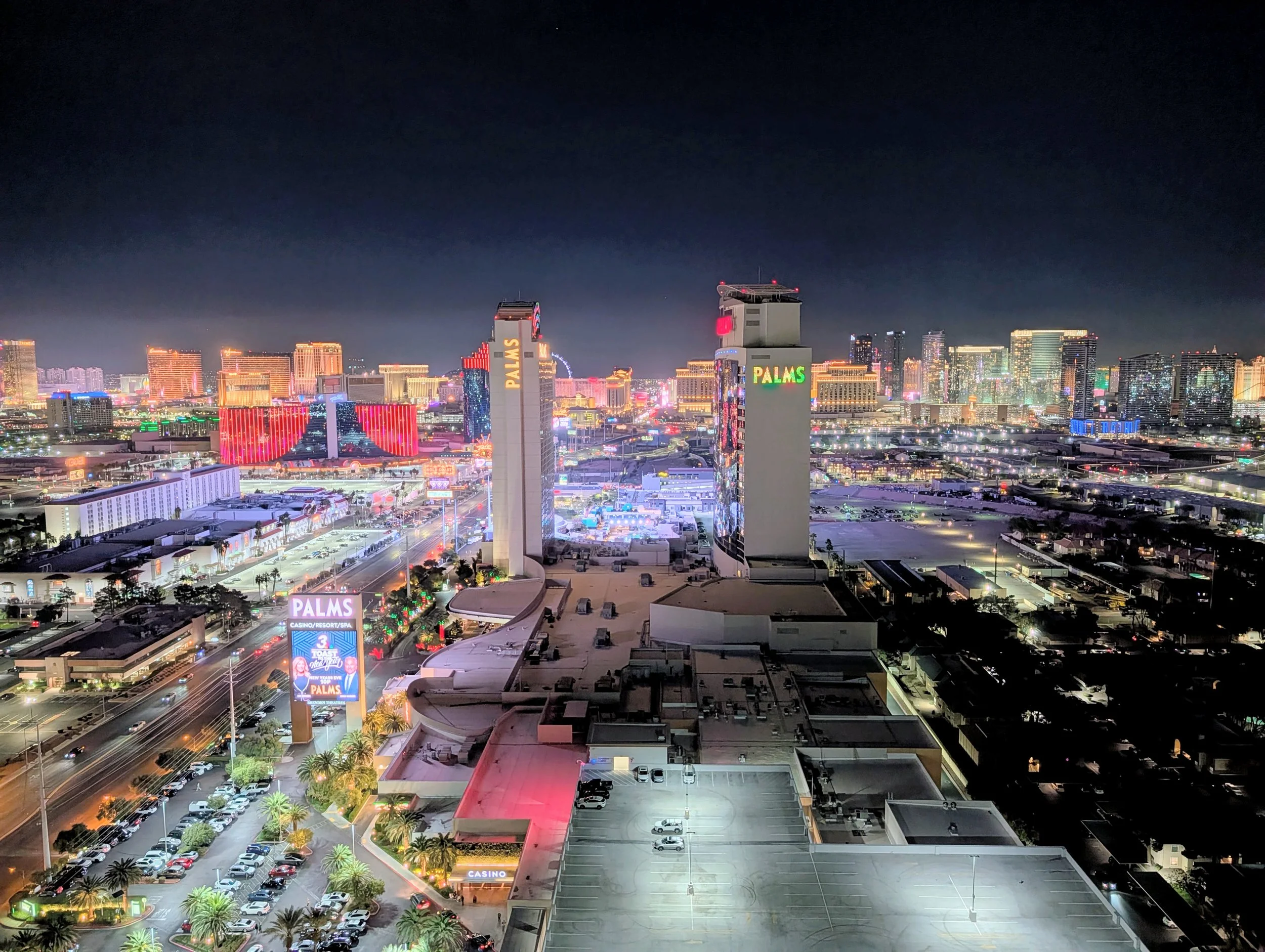 Nighttime aerial view of Las Vegas strip with numerous high-rise hotels and casinos illuminated with colorful lights, featuring the Palms hotel and casino with its tall towers and bright signage.