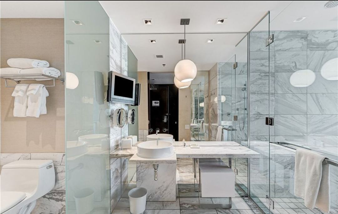 Modern hotel bathroom with marble walls and floors, glass shower enclosure, double sinks, wall-mounted TV, and plush towels.