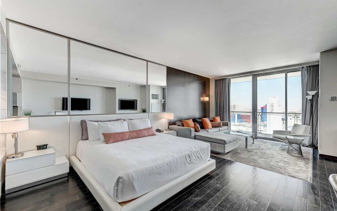 Modern hotel room with a large bed, a sitting area with a gray sofa and pillows, and floor-to-ceiling windows showing a city view.