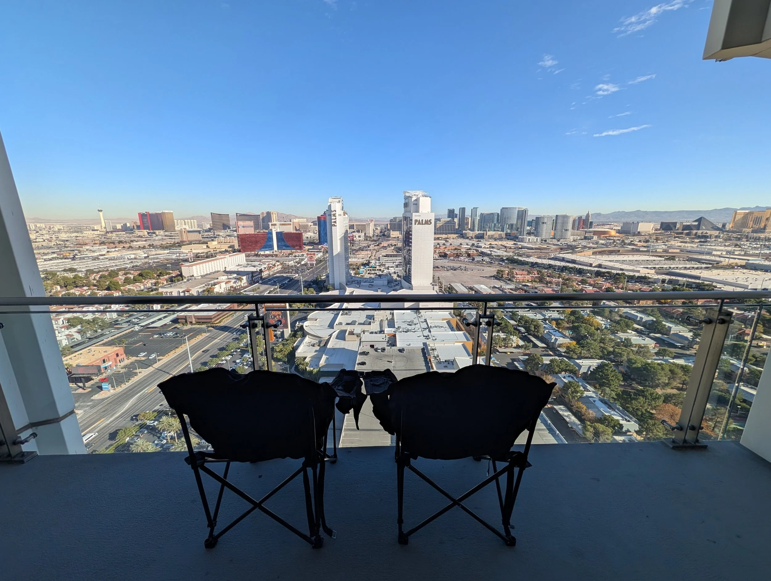 Two black folding chairs on a balcony with a glass railing, overlooking the Las Vegas skyline and desert landscape.