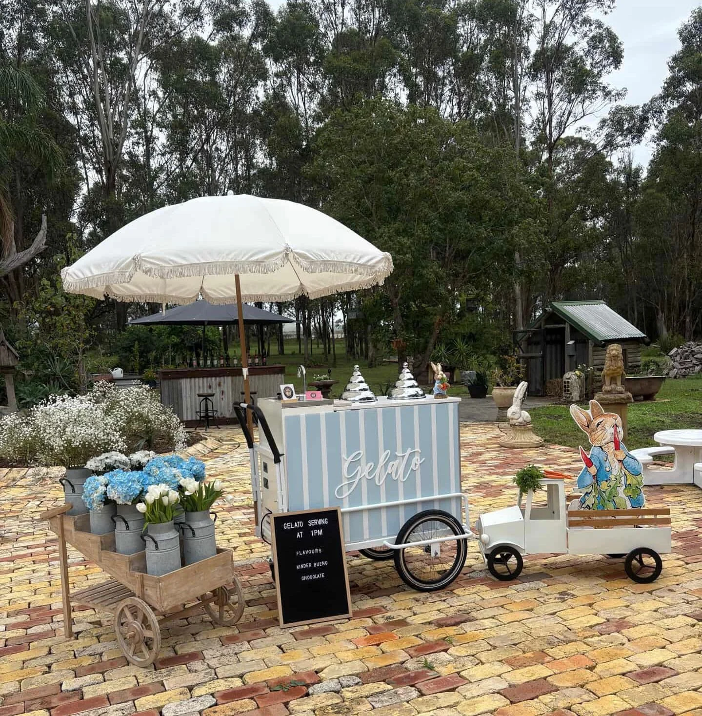 Outdoor gelato cart with a white umbrella, decorated with flowers and a sign, set on a brick patio with trees in the background.
