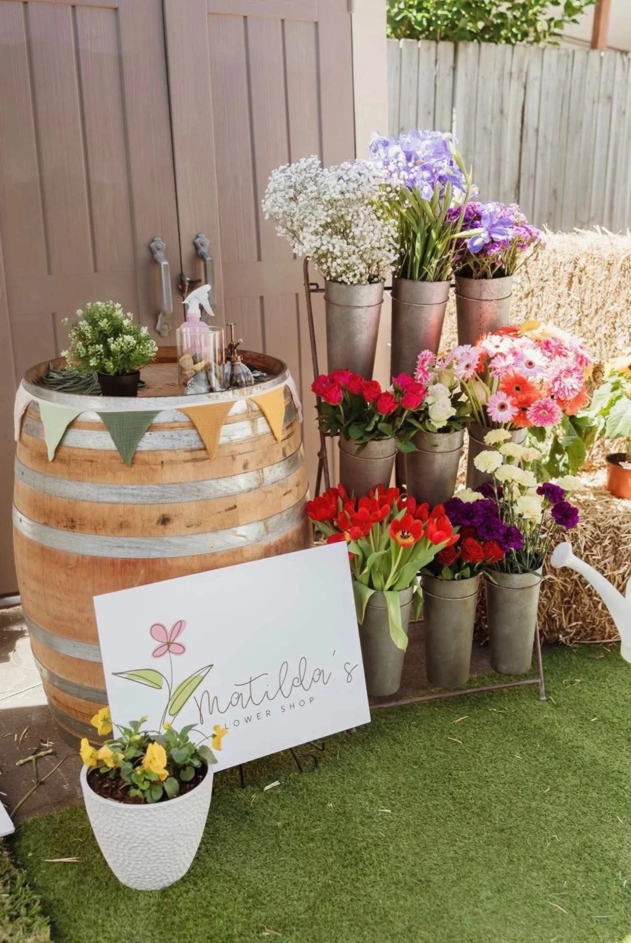 A display of colorful flowers in metal pots on a stand, next to a wooden barrel with a banner, a small potted plant, and a sign for Matilda's Flower Shop, with a watering can and hay bale in the background.