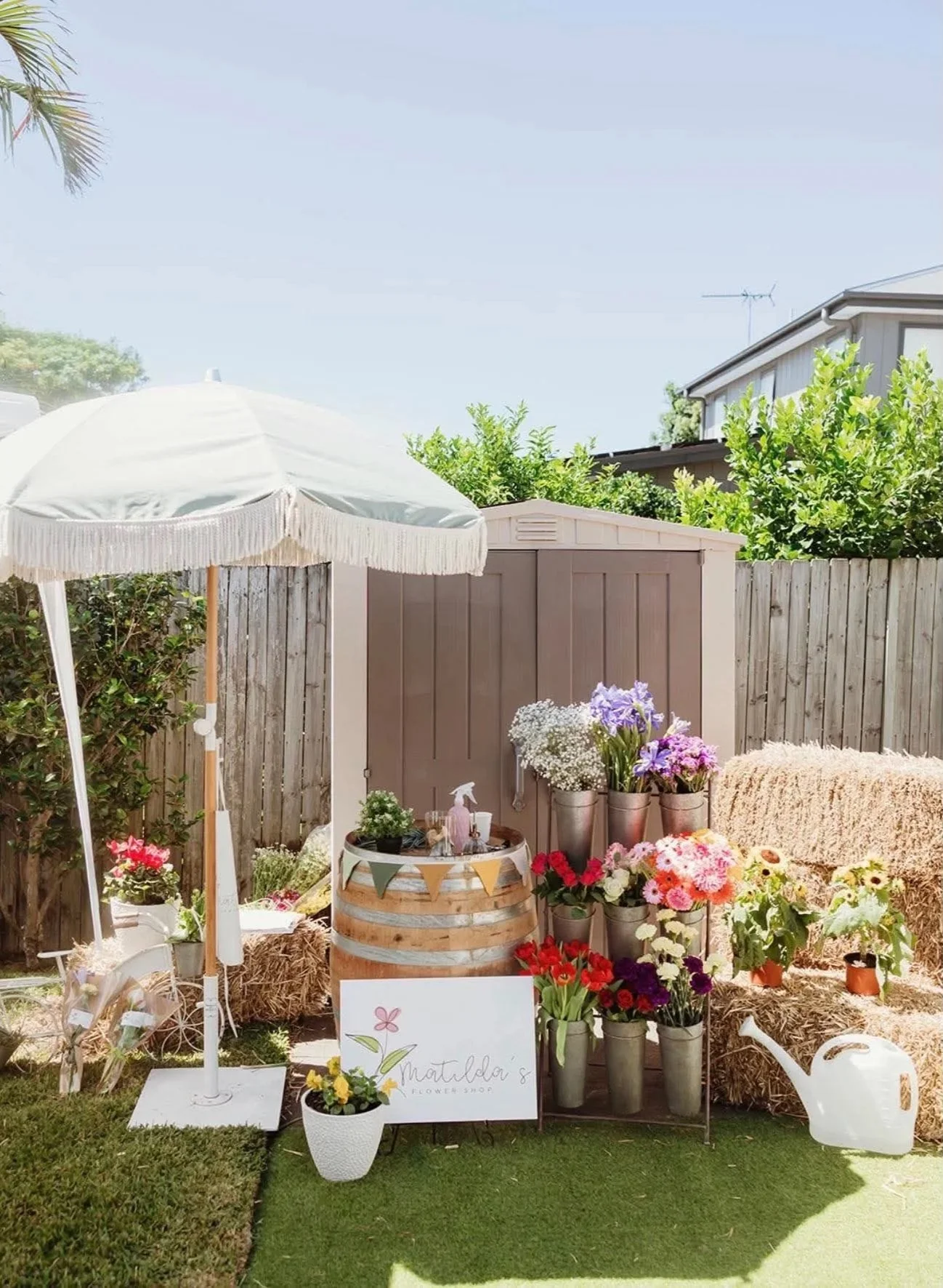 A garden setup featuring a white parasol, a white flower cart, colorful flowers in metal buckets, a hay bale, a white watering can, and a sign reading 'Matilda's Flower Shop', all arranged on artificial grass with a wooden fence and backyard in the b
