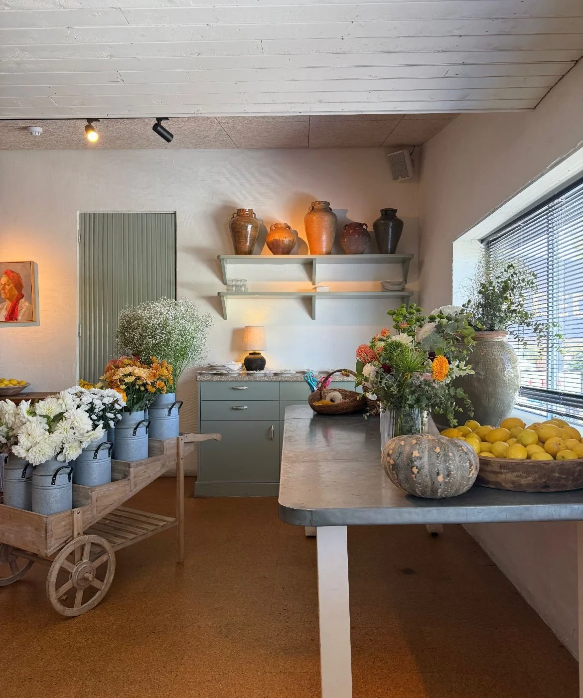 Interior of a cozy room with a light-colored ceiling, a window with blinds, and various decorated vases, flowers, and cookbooks on shelves and tables.