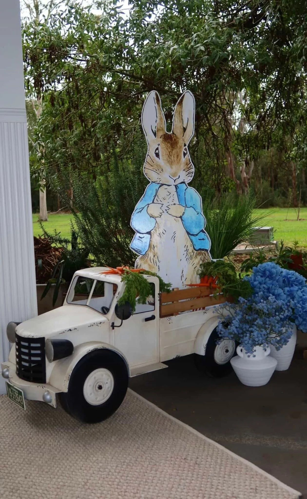 Decorative display featuring a vintage truck with a large bunny figure wearing a blue jacket, surrounded by flowers and greenery.