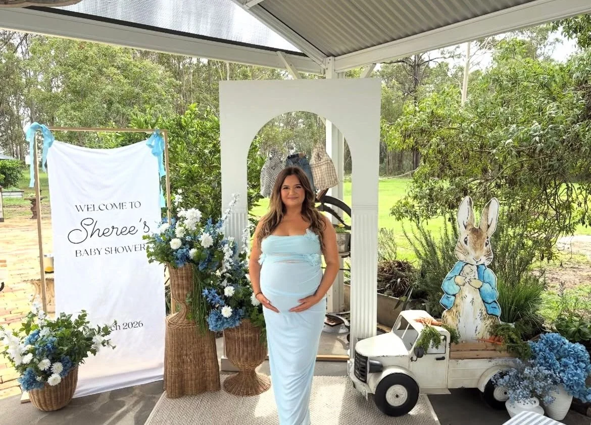 Pregnant woman in a light blue dress at baby shower with floral arrangements, a welcome sign, a white toy truck, and a bunny cutout in an outdoor setting.
