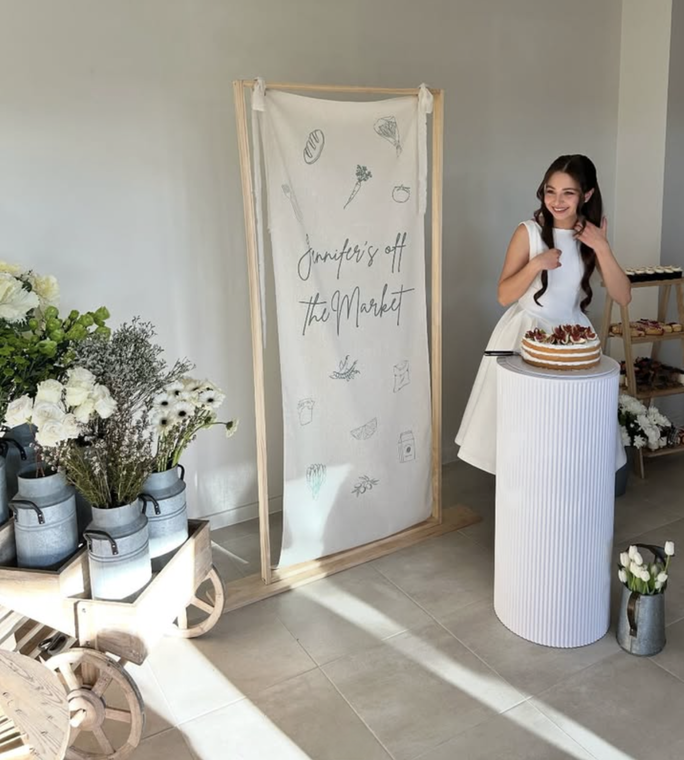 A young woman in a white dress smiling next to a birthday cake on a white, ribbed pedestal at an event. Behind her, a sign reads 'Jennifer's off the Market' and is decorated with sketches of food items and plants. To her left, there is a cart with vases of white and green flowers.