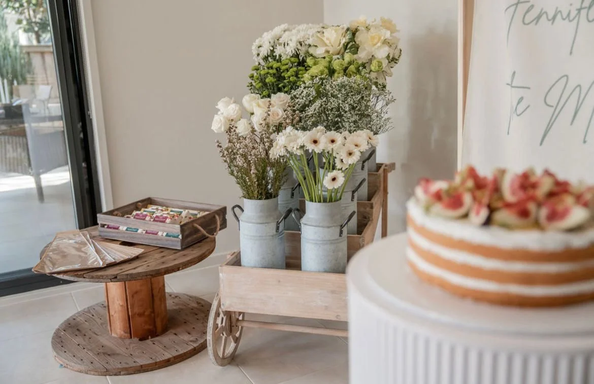 A wooden table with a cardboard box and paper bag on it, a shopping cart with three metal milk cans filled with white flowers, and a cake with pink flowers in the foreground.