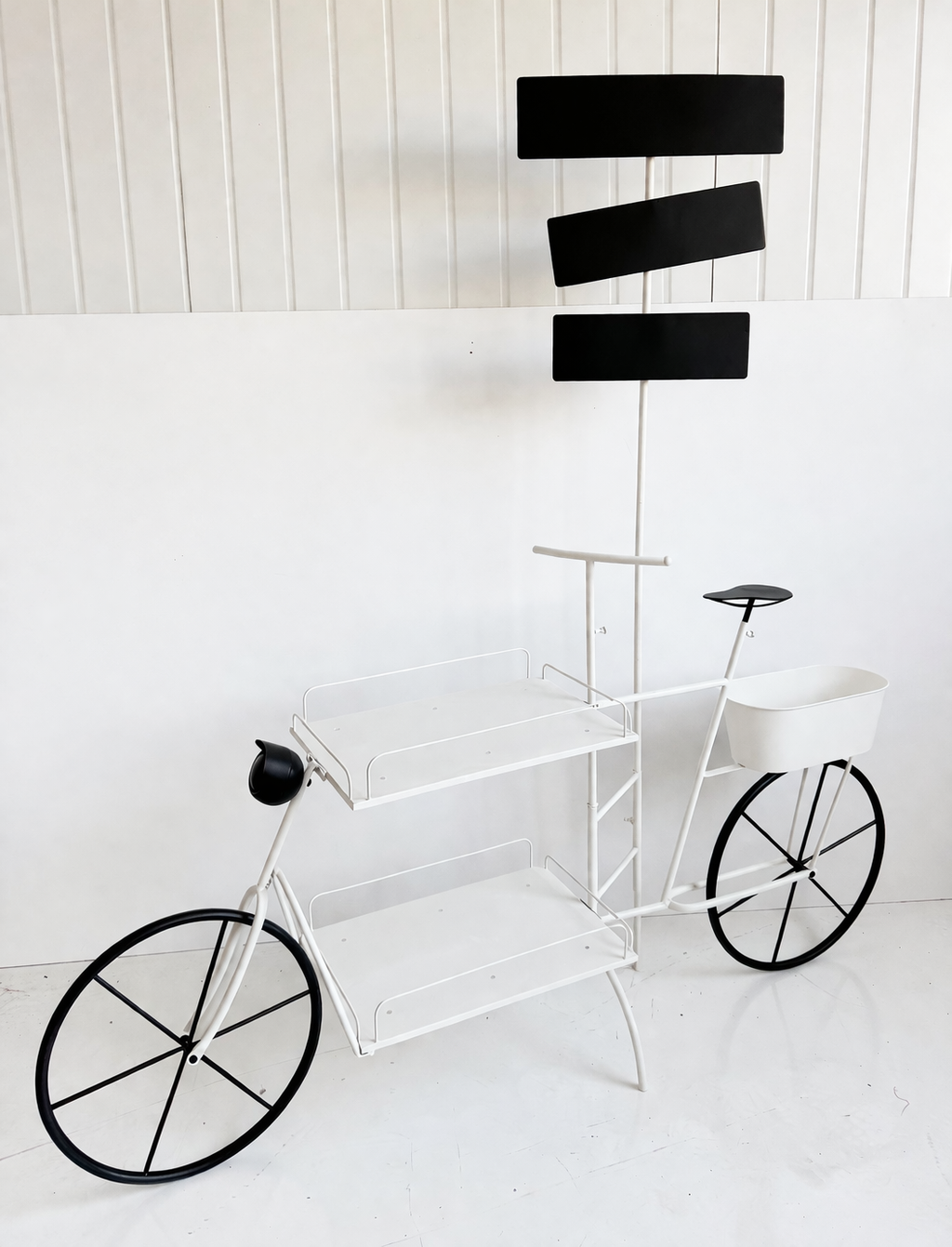 A decorative bicycle-shaped shelf with black rectangular signs on the front and a white basket on the back, standing against a white wall.