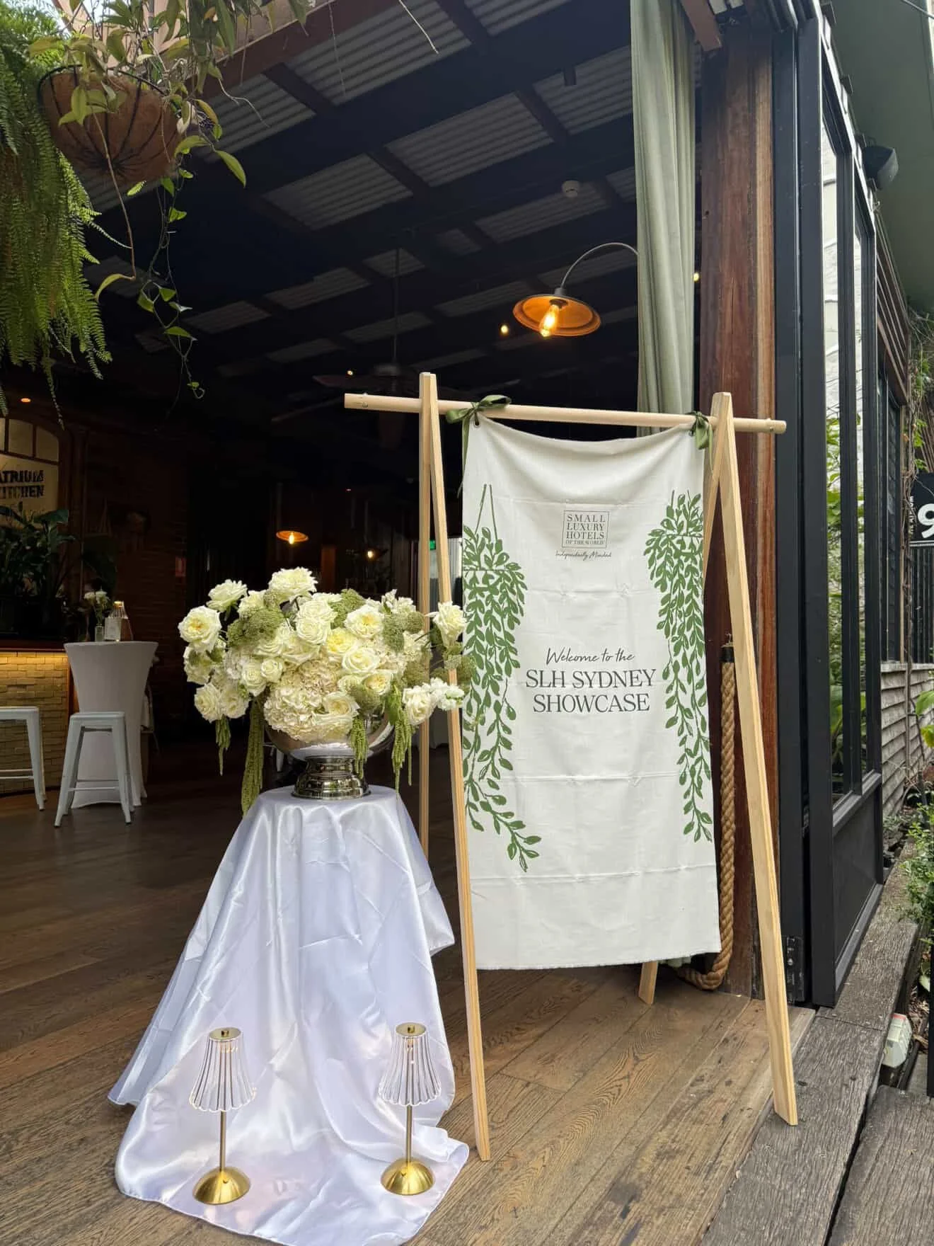 Entrance decorated for the SLH Sydney Showcase with a white floral arrangement on a table covered with a white cloth, and a sign with green foliage design leaning on a wooden stand.