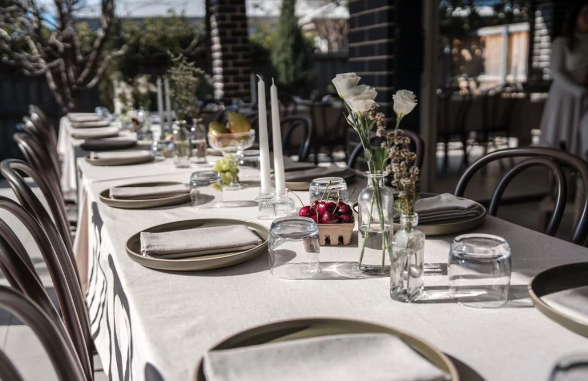 Outdoor dining table set with plates, napkins, glasses, candles, and vases with flowers arranged for a meal.