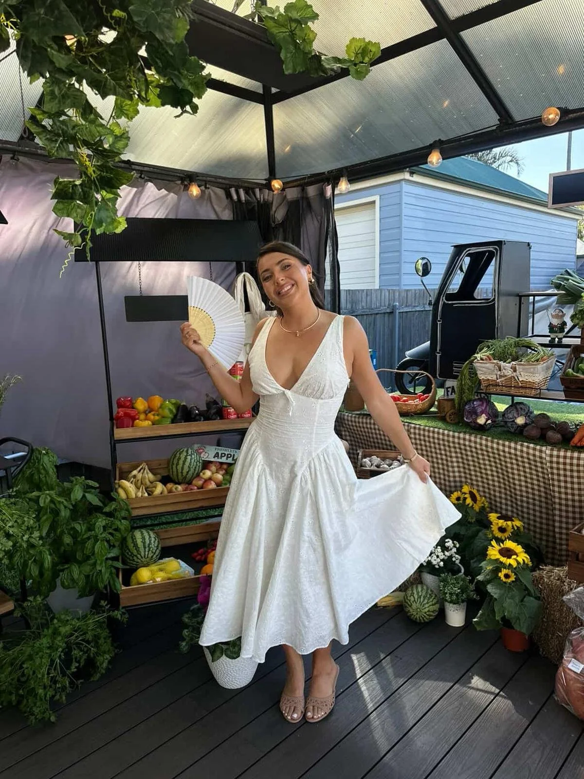 A young woman in a white dress holding a folding fan, standing at an outdoor farmers market stand with fresh produce and flowers, smiling at the camera.