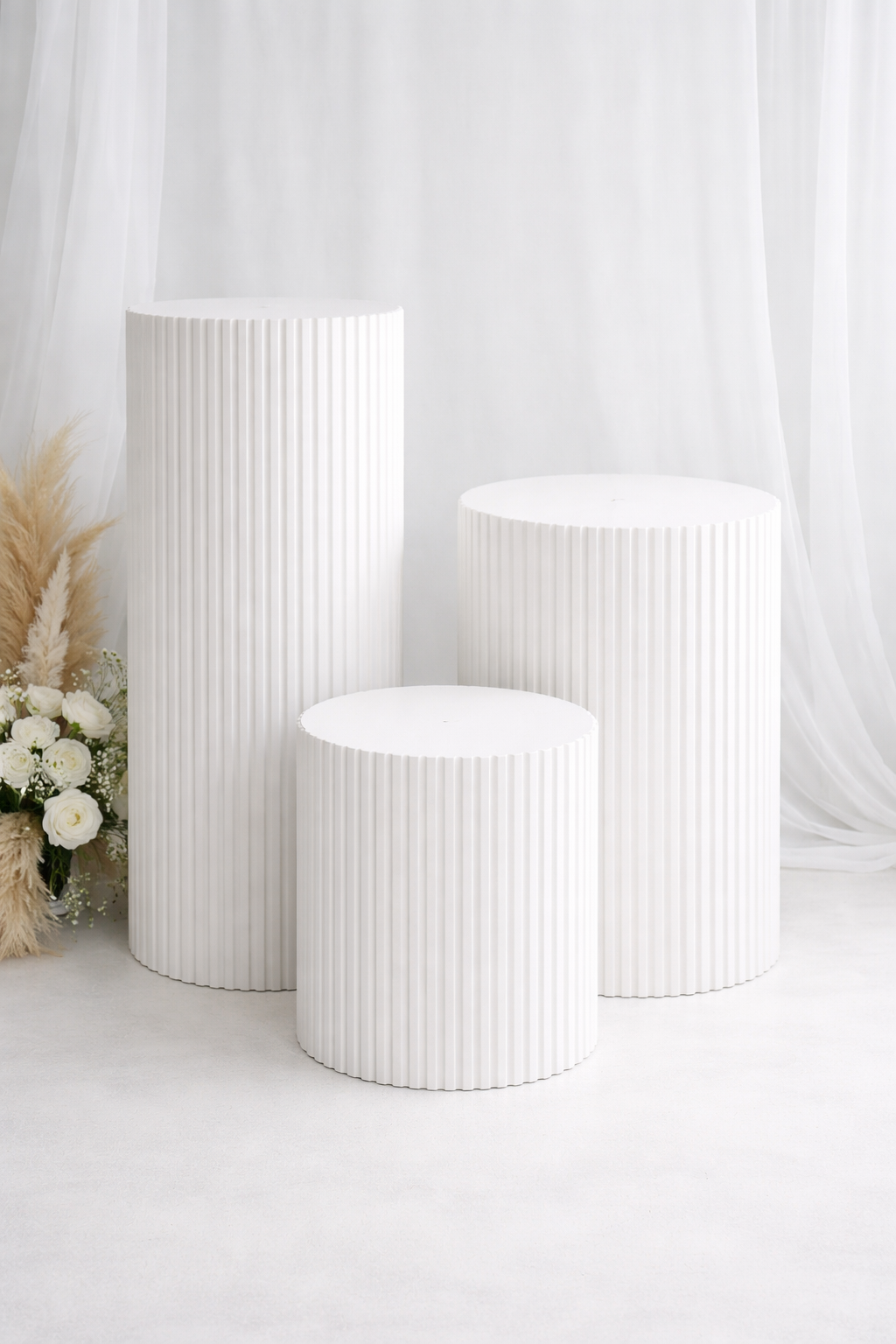 Three white cylindrical pedestals with ribbed texture, arranged in a staggered formation in front of a white backdrop. There are white flowers and beige pampas grass on the left side.