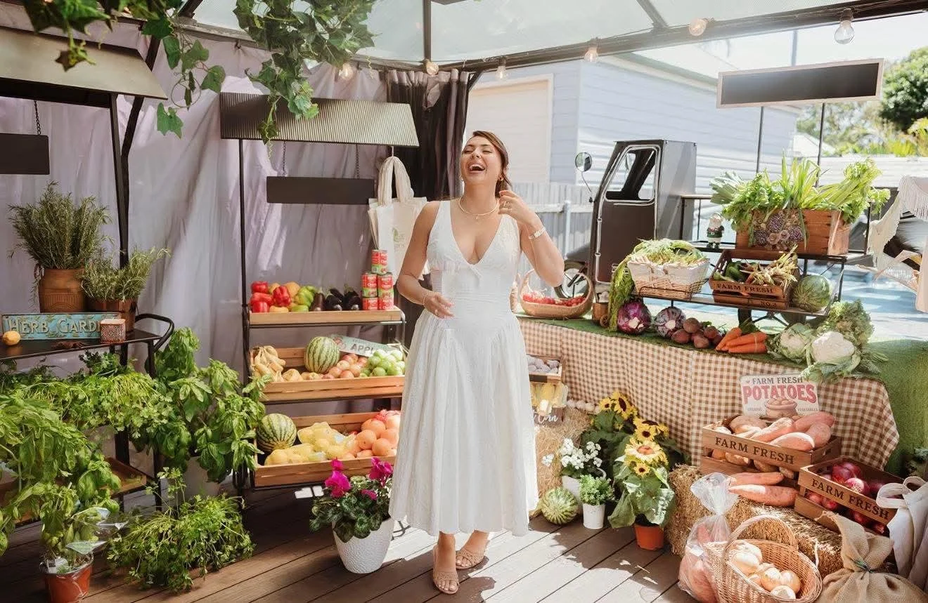 Woman in a white dress laughing at a farmers market stall with fresh vegetables and flowers.
