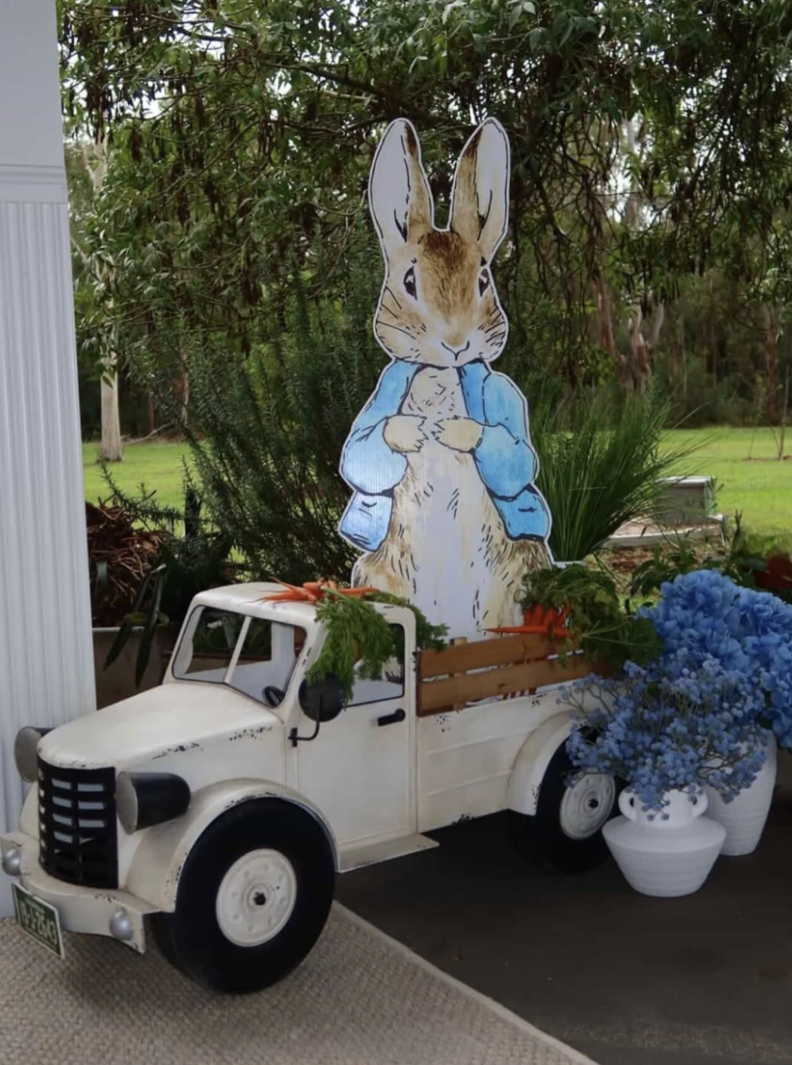 Decorative scene with a vintage white toy truck filled with blue hydrangeas and greenery, a large illustrated rabbit figure standing behind the truck wearing a blue jacket, set against a background of trees and plants.