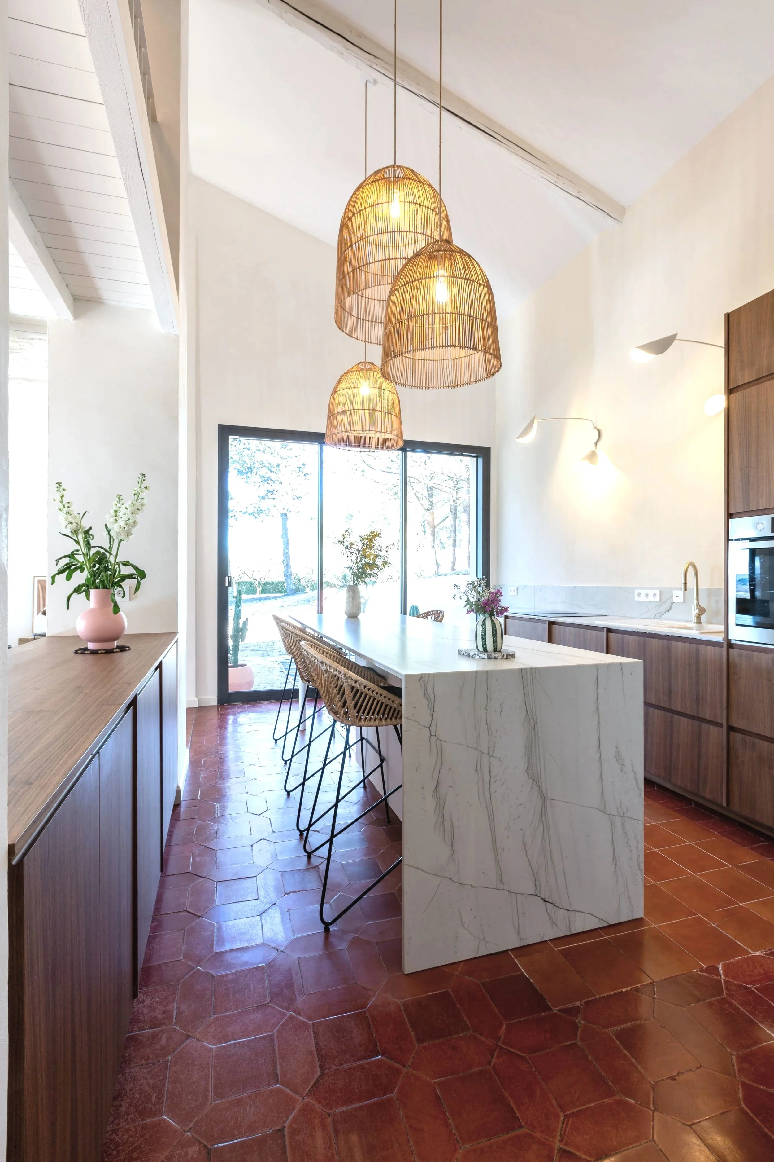 Une cuisine lumineuse  en facade bois noyer plum living, un plan de travail en marbre blanc, des chaises en osier, des luminaires suspendus en rotin, des lampes murales blanches, réalisé par Laure Galippe , architecte d'intérieur à Aix-en-Provence