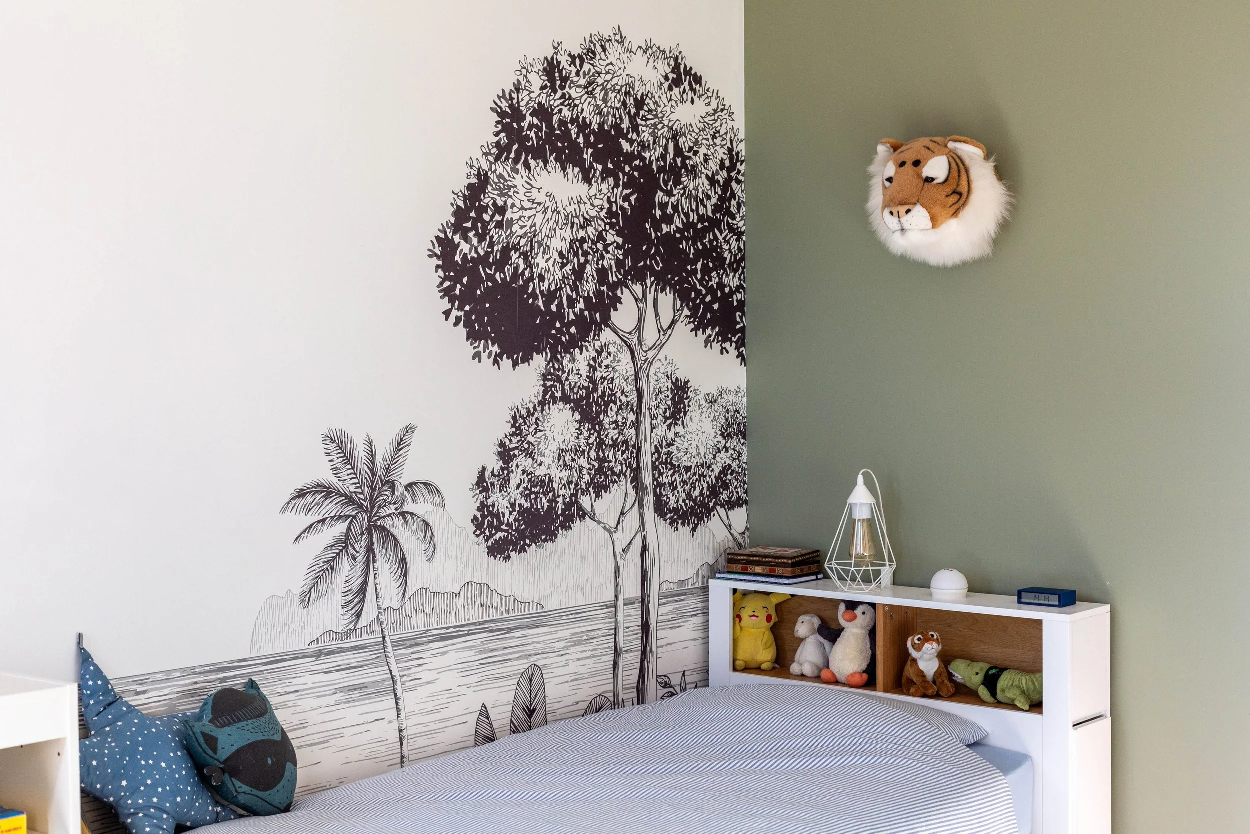 Chambre d'enfant avec un mur décoré d'une fresque de paysages avec des arbres et une plage, un lit avec des coussins et une tête de lit représentant un animal, un dormeur avec des jouets et des livres, une lampe Laure galippe rénovation eguilles