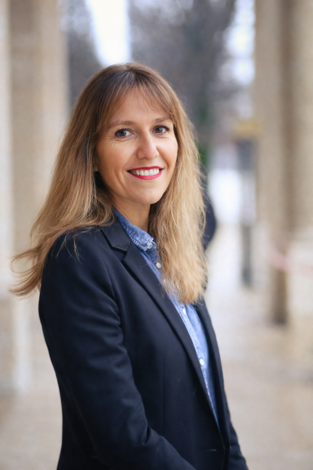 Laure Galippe , architecte d'intérieur à Aix-en-Provence souriante portant un blazer noir et une chemise bleue, debout dans un couloir architectural avec colonnes.